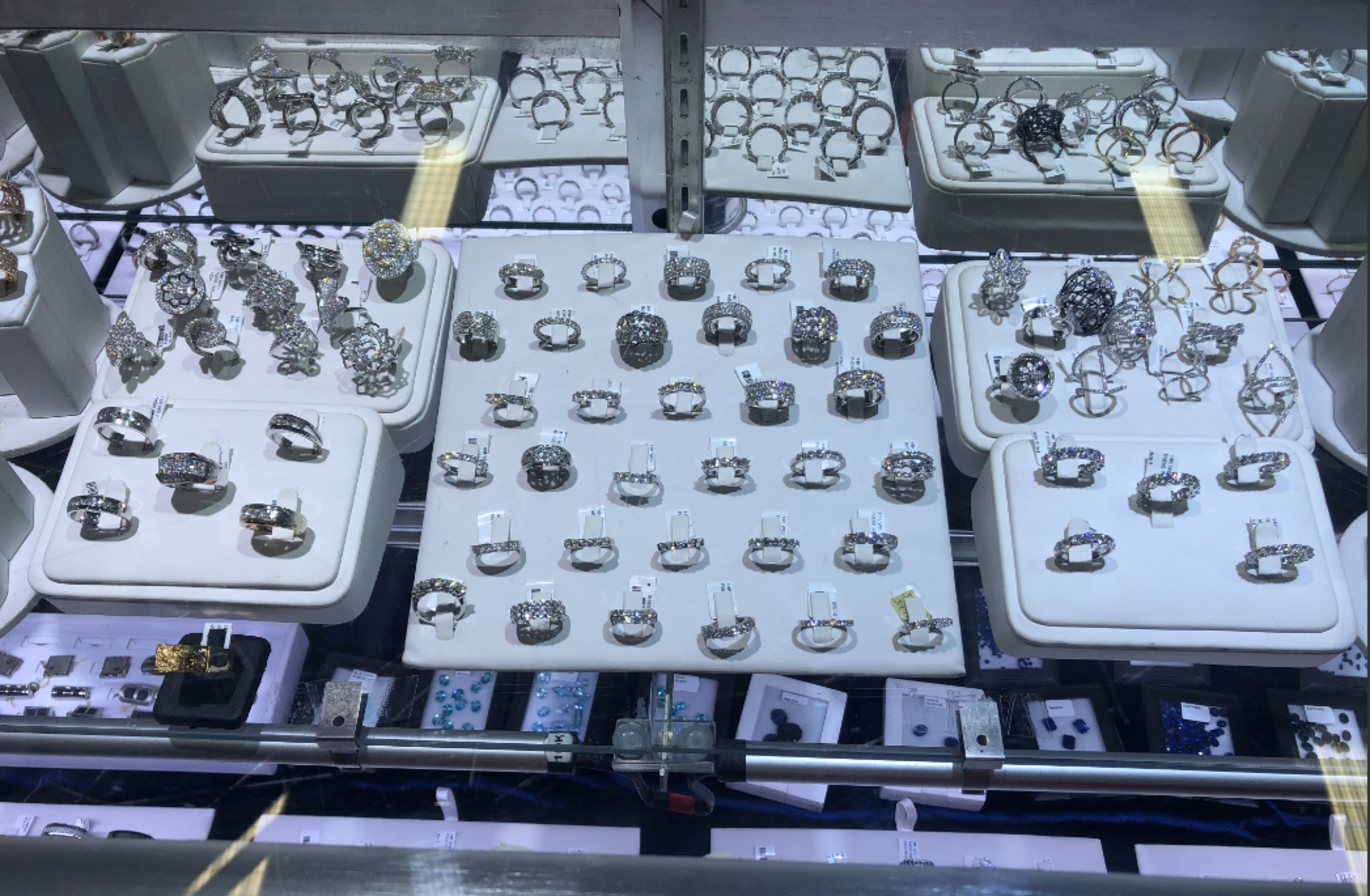 The image shows a display case filled with a variety of rings, including many with intricate designs and sparkling stones, arranged neatly on white platforms.