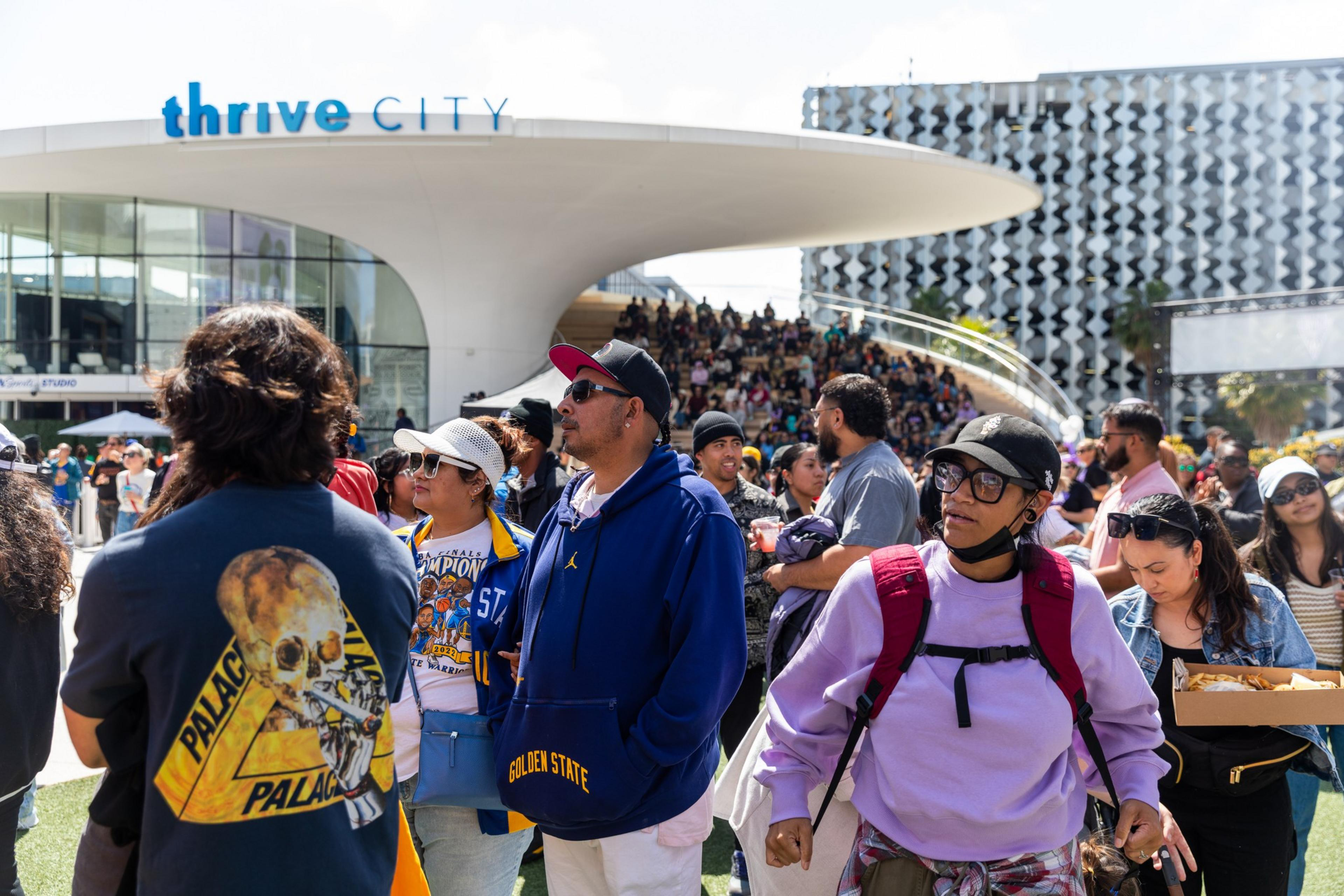 A diverse crowd gathers under a sunny sky at Thrive City, with a modern building in the background. Some individuals are attentive, others converse or eat.