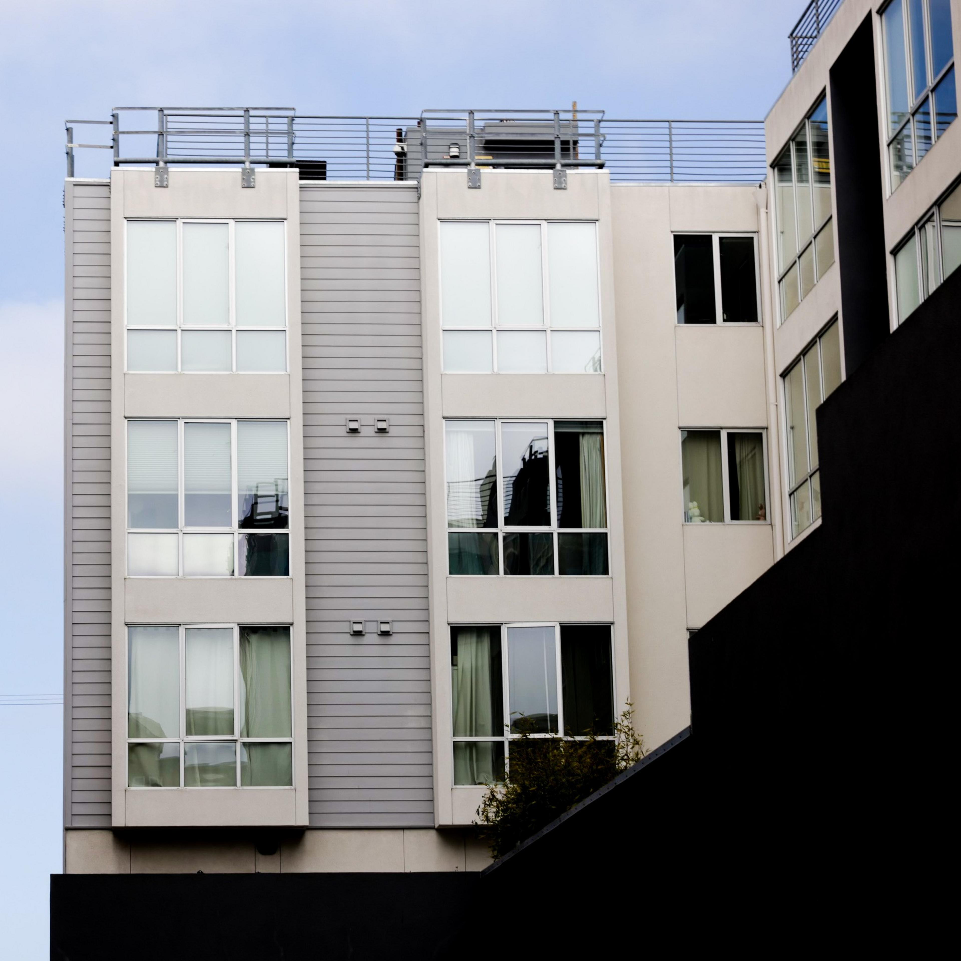 The image shows a modern, multi-story residential building with large windows and a rooftop railing under a partially cloudy sky.