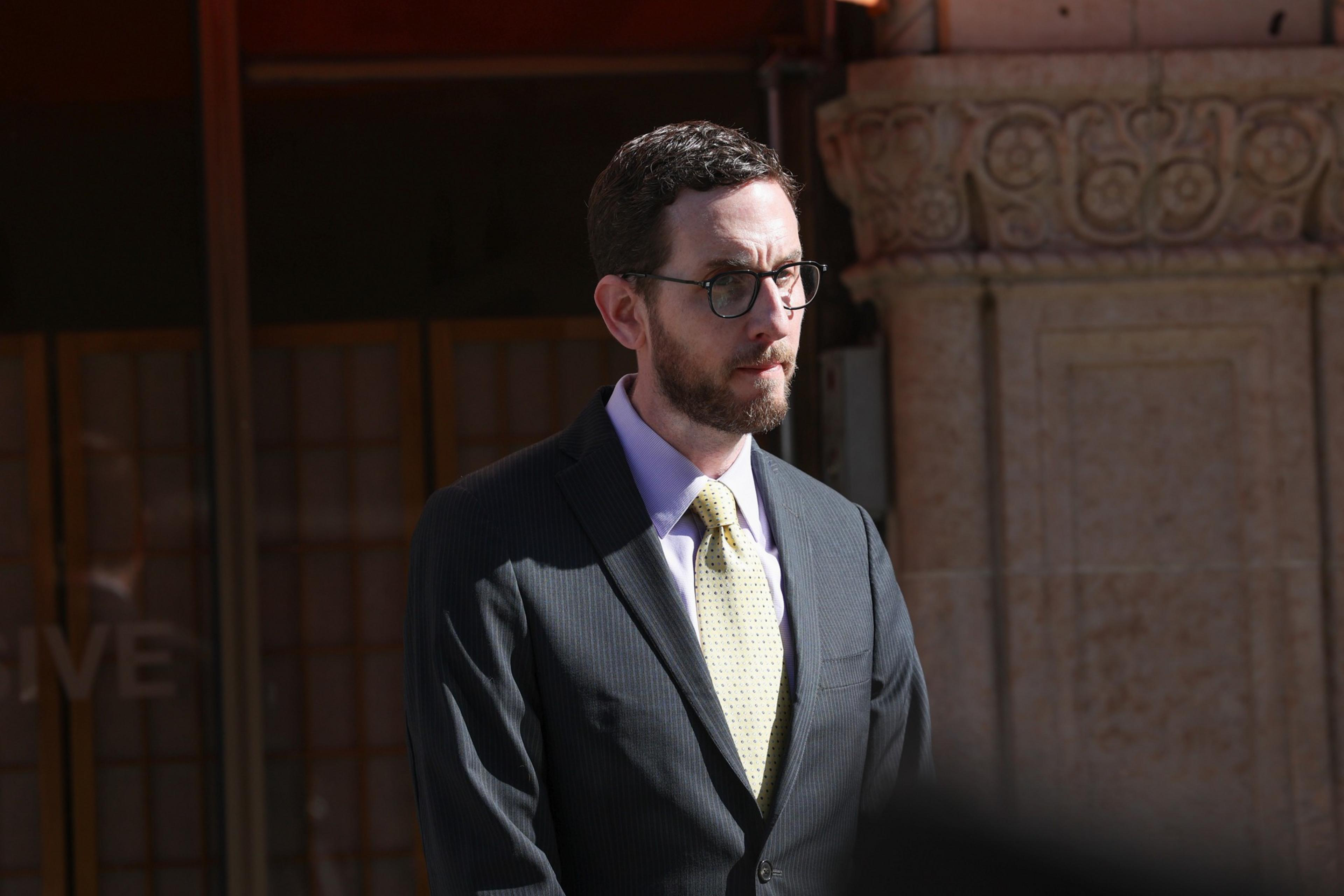 A man with glasses and a beard wears a dark suit, light yellow tie, and light purple shirt, standing near a stone building in sunlight.