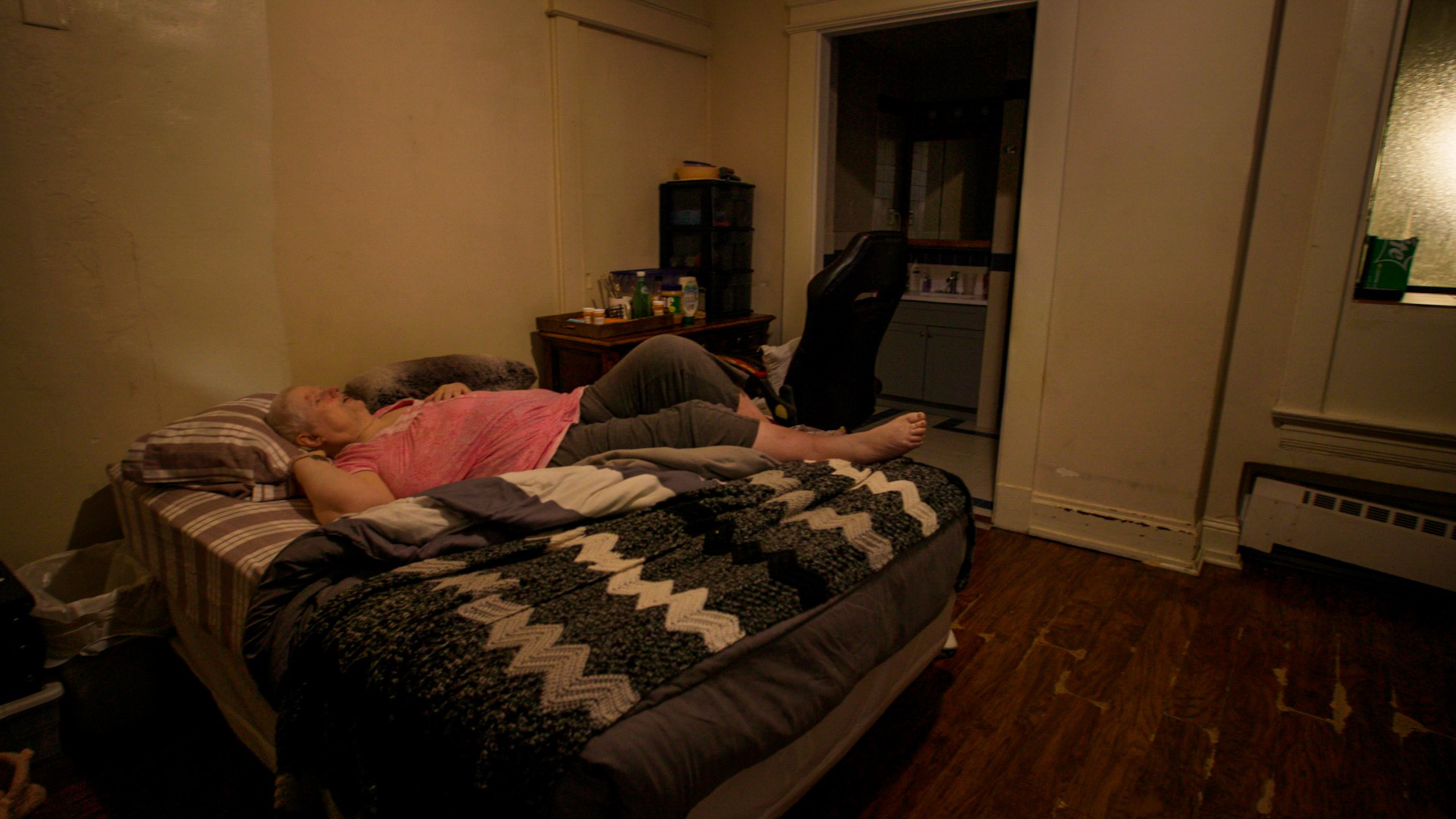 An elderly person lies on a striped bed covered partly with a black-and-white patterned blanket, in a dimly lit room with wooden floors and sparse furnishings.