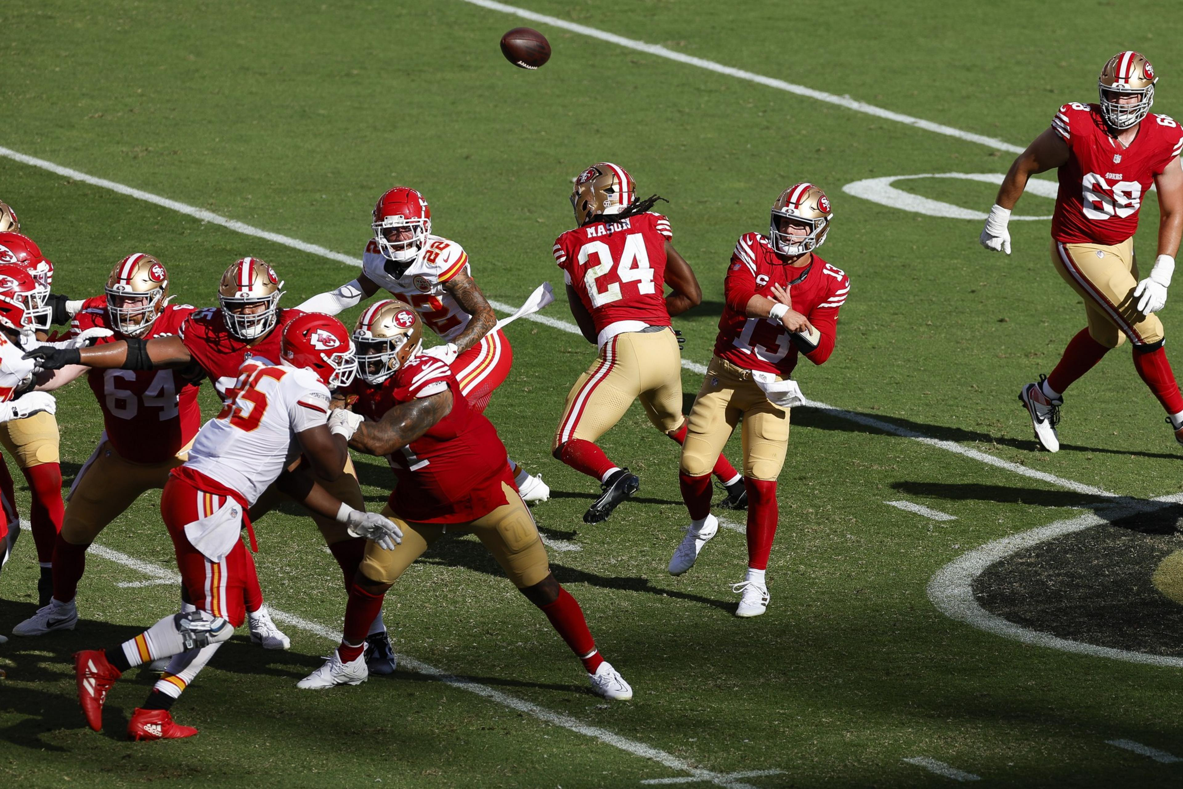 A football player in a red and gold uniform throws a pass while other players in red and white positions around him attempt to block or defend.