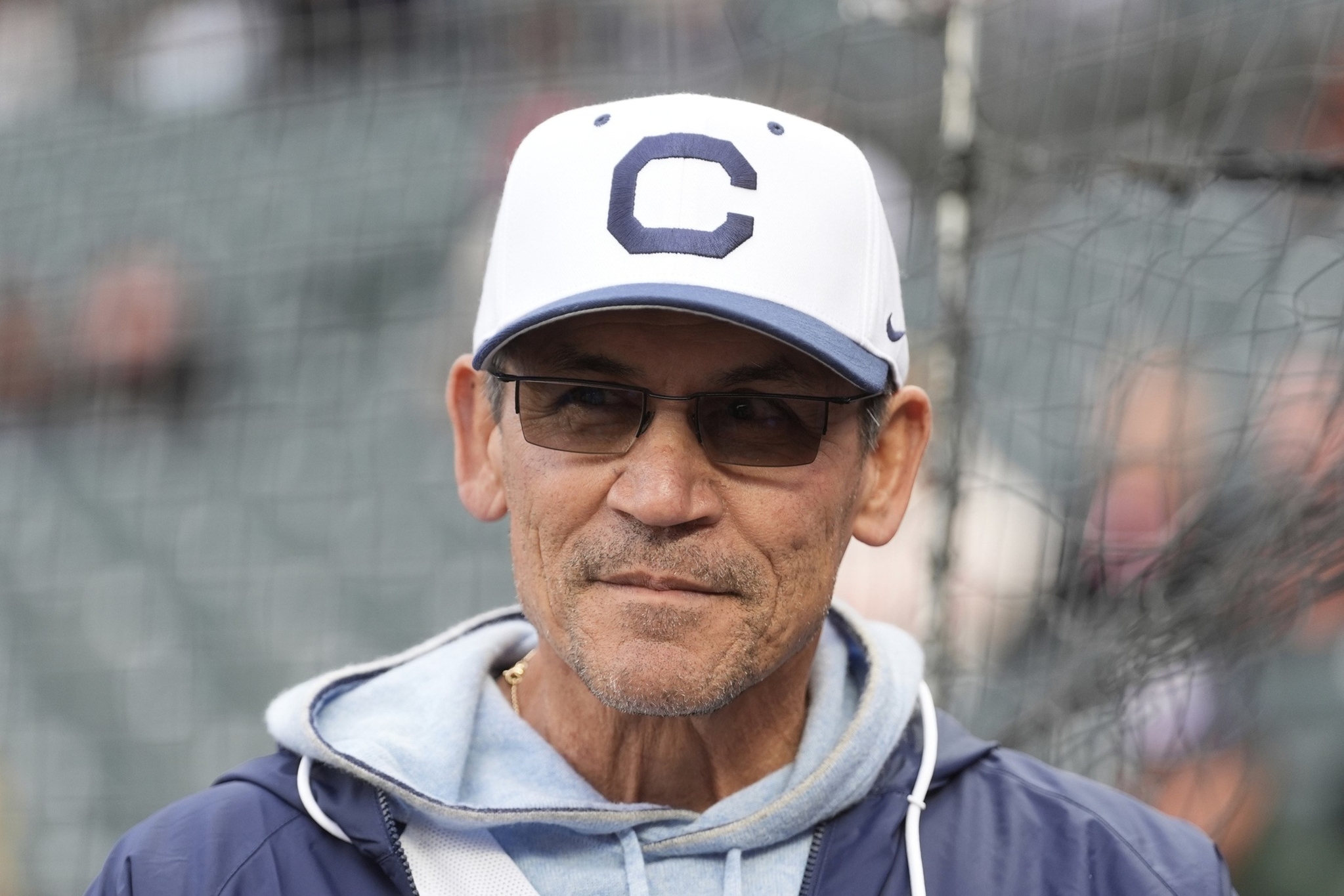 A middle-aged man wears dark sunglasses, a white baseball cap with a blue "C," a light blue hoodie, and a dark blue jacket, standing against a netted background.