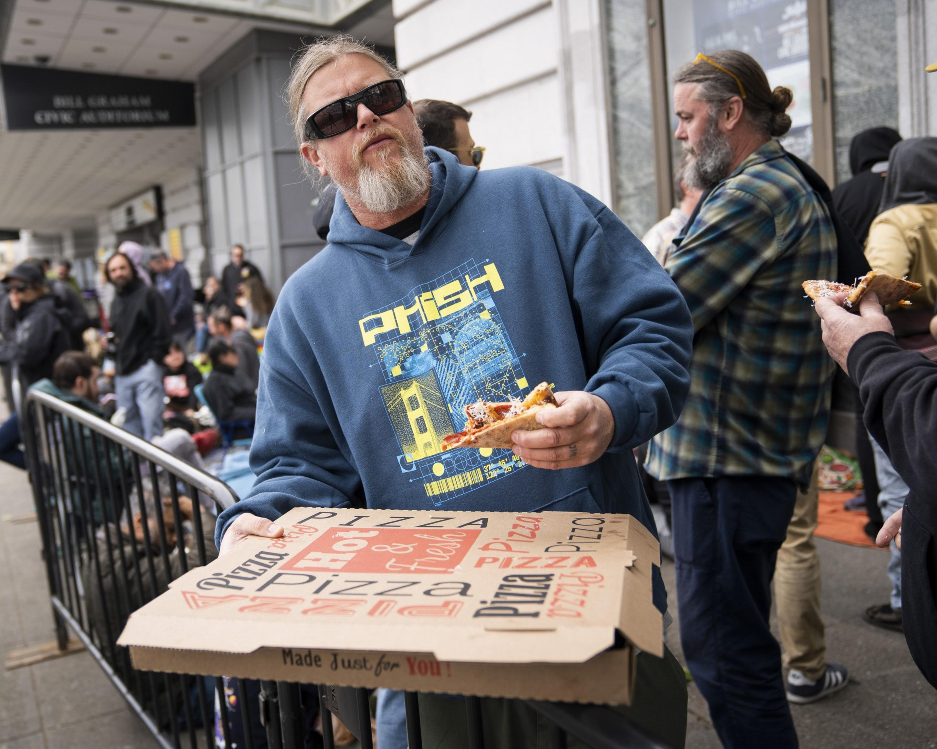 A bearded man in sunglasses and a blue hoodie holds a pizza slice near a pizza box. People are gathered outside a building with a &quot;Civic Auditorium&quot; sign.