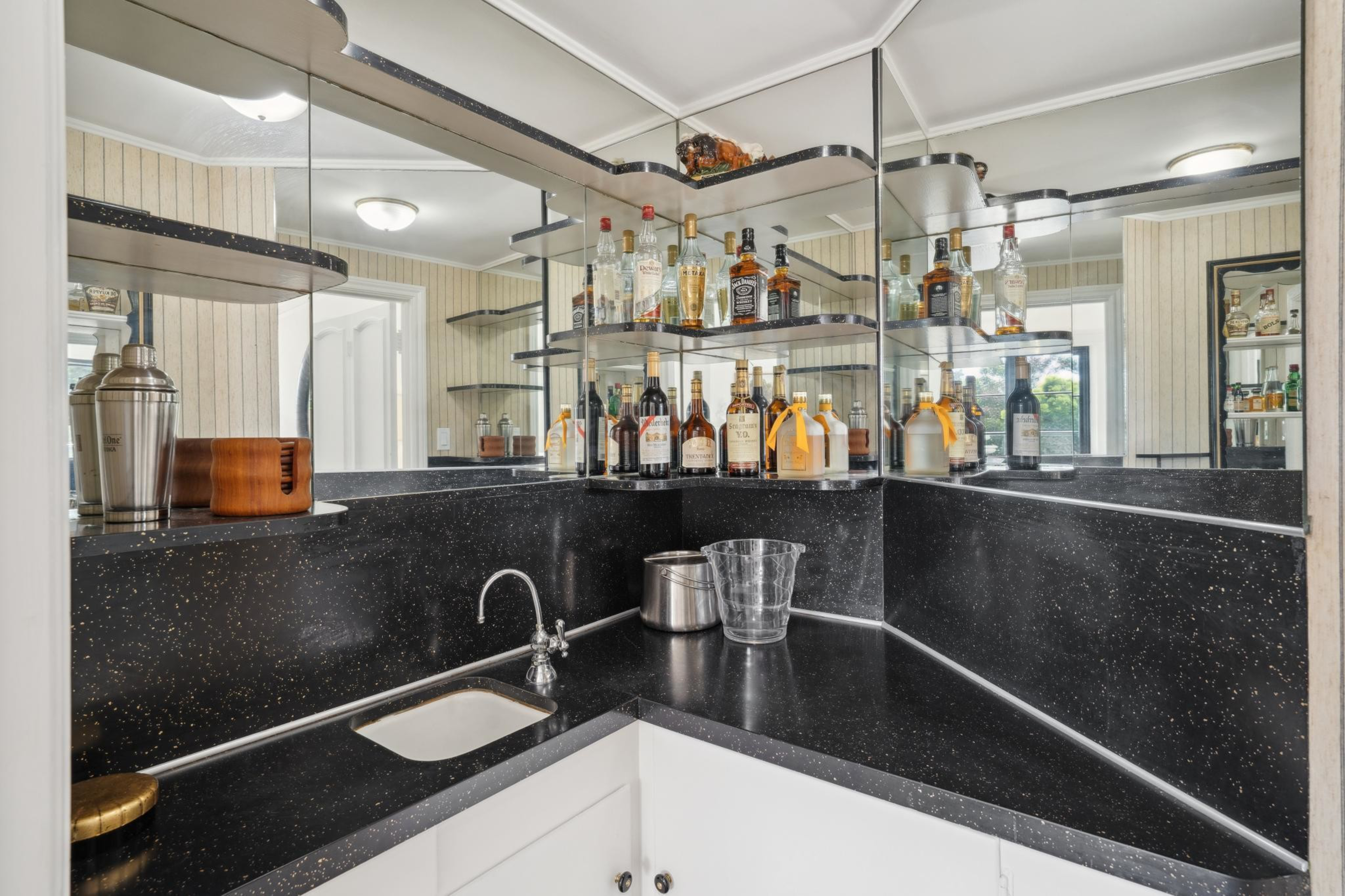 A home bar corner with a black speckled countertop, a small sink, shelves filled with liquor bottles, mirrored walls, an ice bucket, and a cocktail shaker.