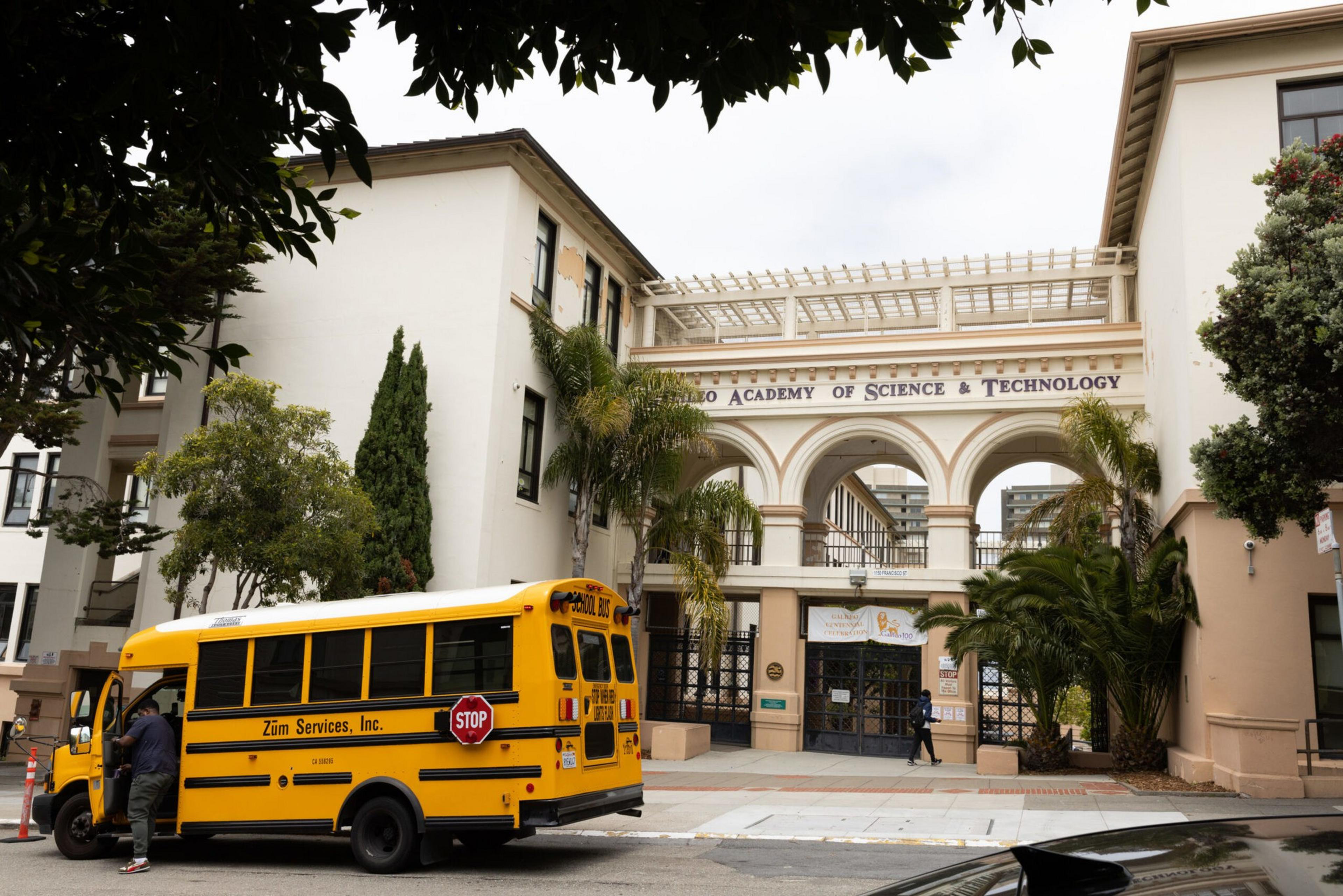 A yellow school bus labeled "Zum Services, Inc." is parked outside the light-colored building of the Galileo Academy of Science & Technology, surrounded by greenery.