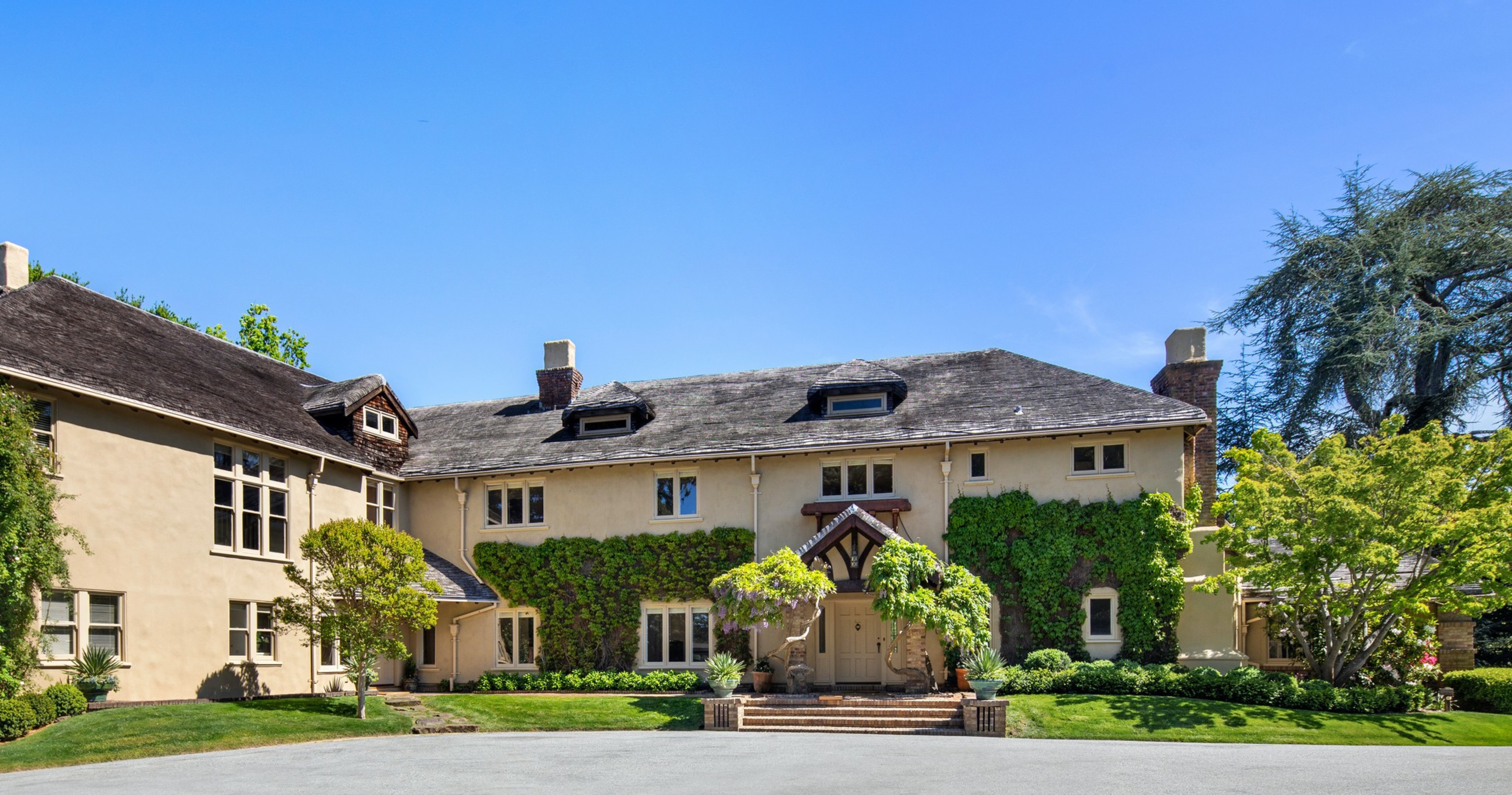 A large beige house with ivy on the front, multiple chimneys, dormer windows, and surrounded by manicured green trees and bushes under a clear blue sky.