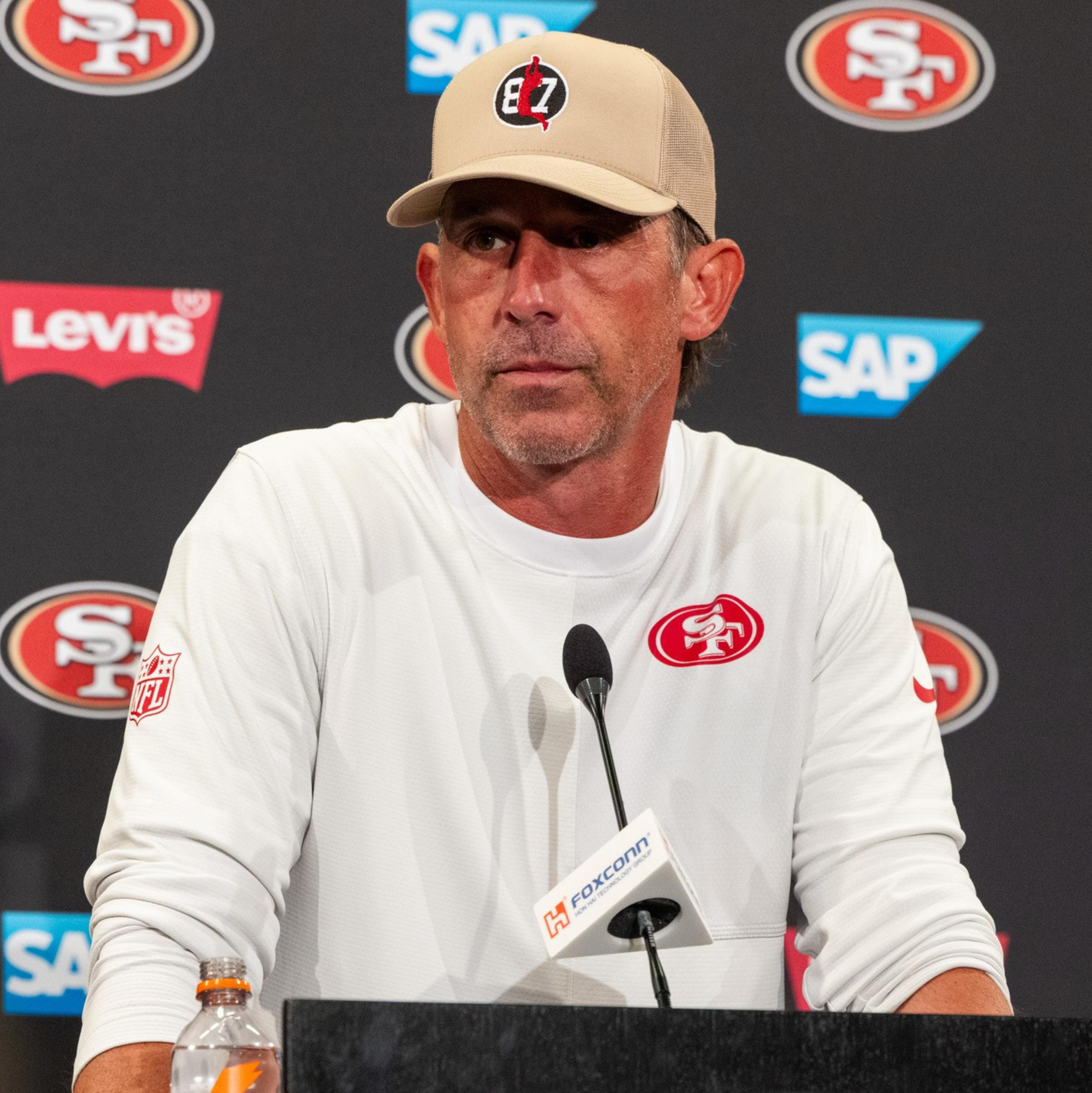 A man wearing a white long-sleeve shirt and beige cap with a logo stands at a podium with a microphone, in front of a backdrop with 49ers and sponsor logos.
