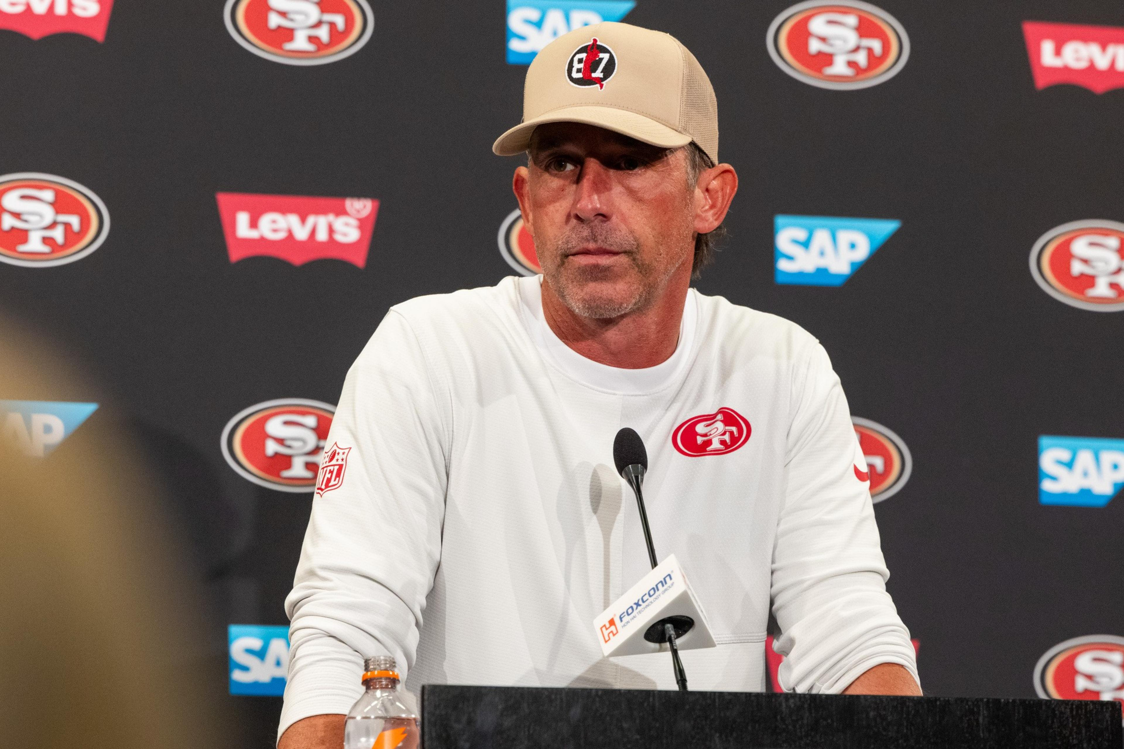 A man wearing a white long-sleeve shirt and beige cap with a logo stands at a podium with a microphone, in front of a backdrop with 49ers and sponsor logos.