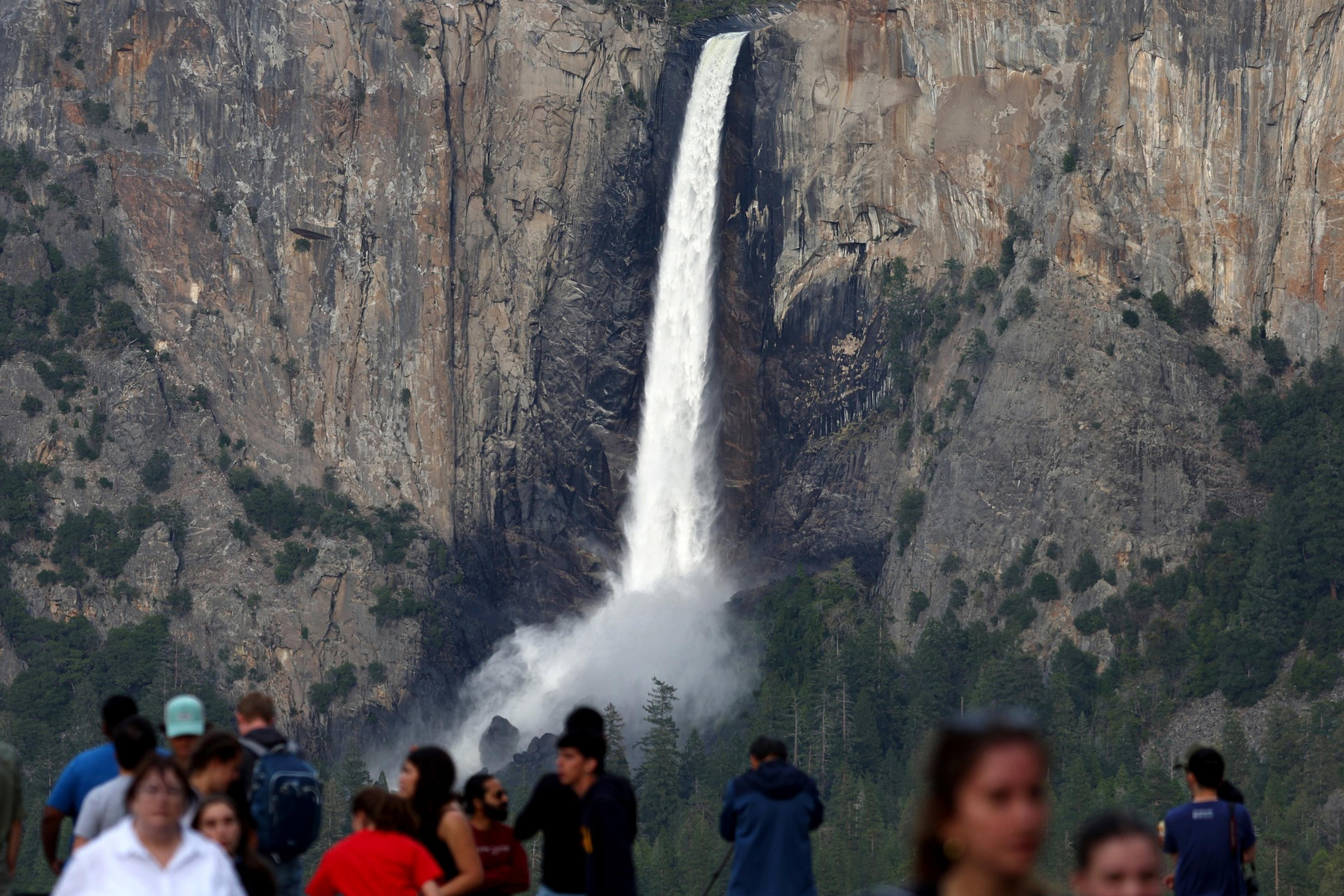 A tall waterfall cascades down a rugged cliffside with mist rising at its base. In the foreground, a crowd of people gathers, partially blurred, observing the scenery.
