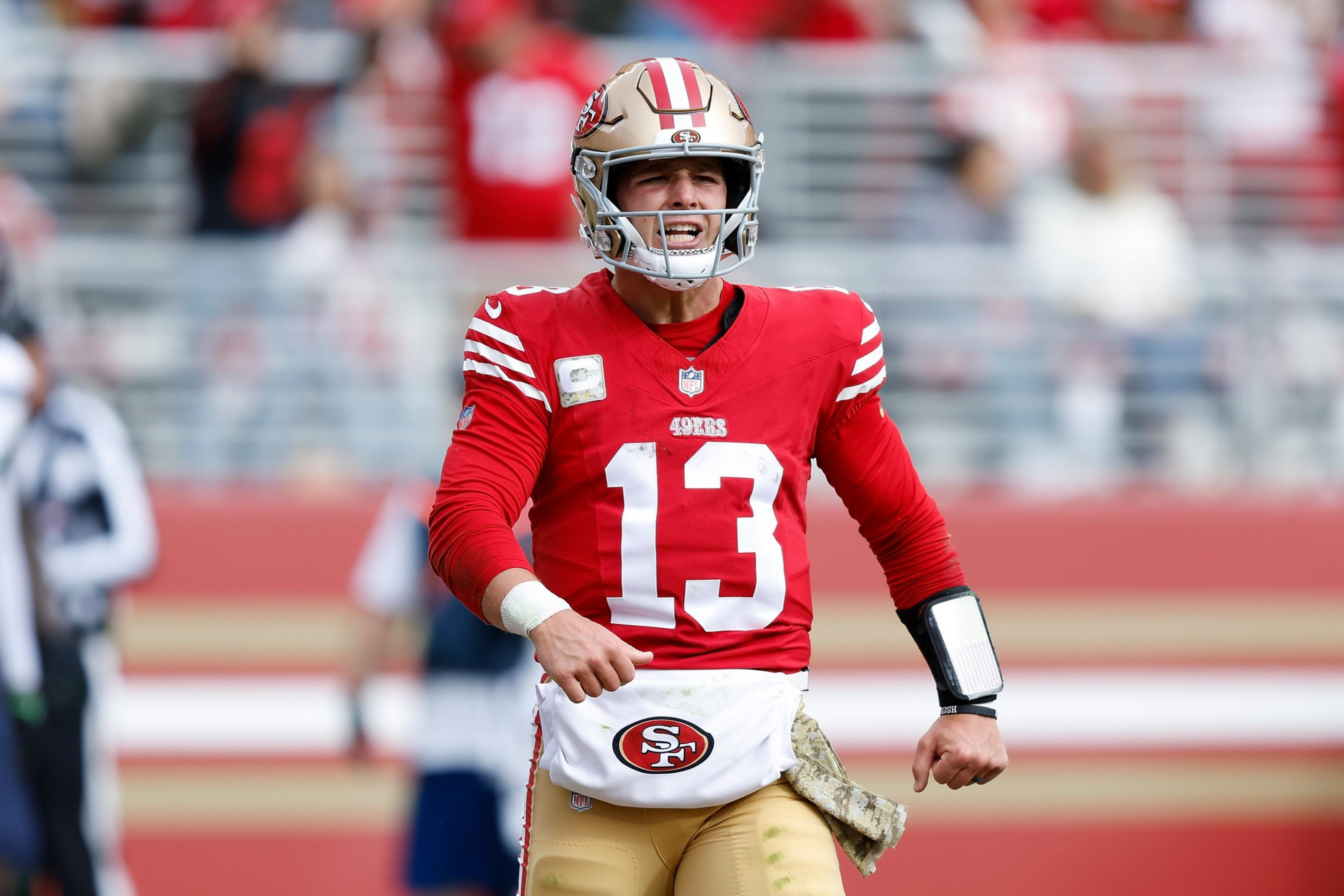 A football player in a red jersey with the number 13 is on the field, wearing a helmet and appearing to be in action, with a background of blurred spectators.
