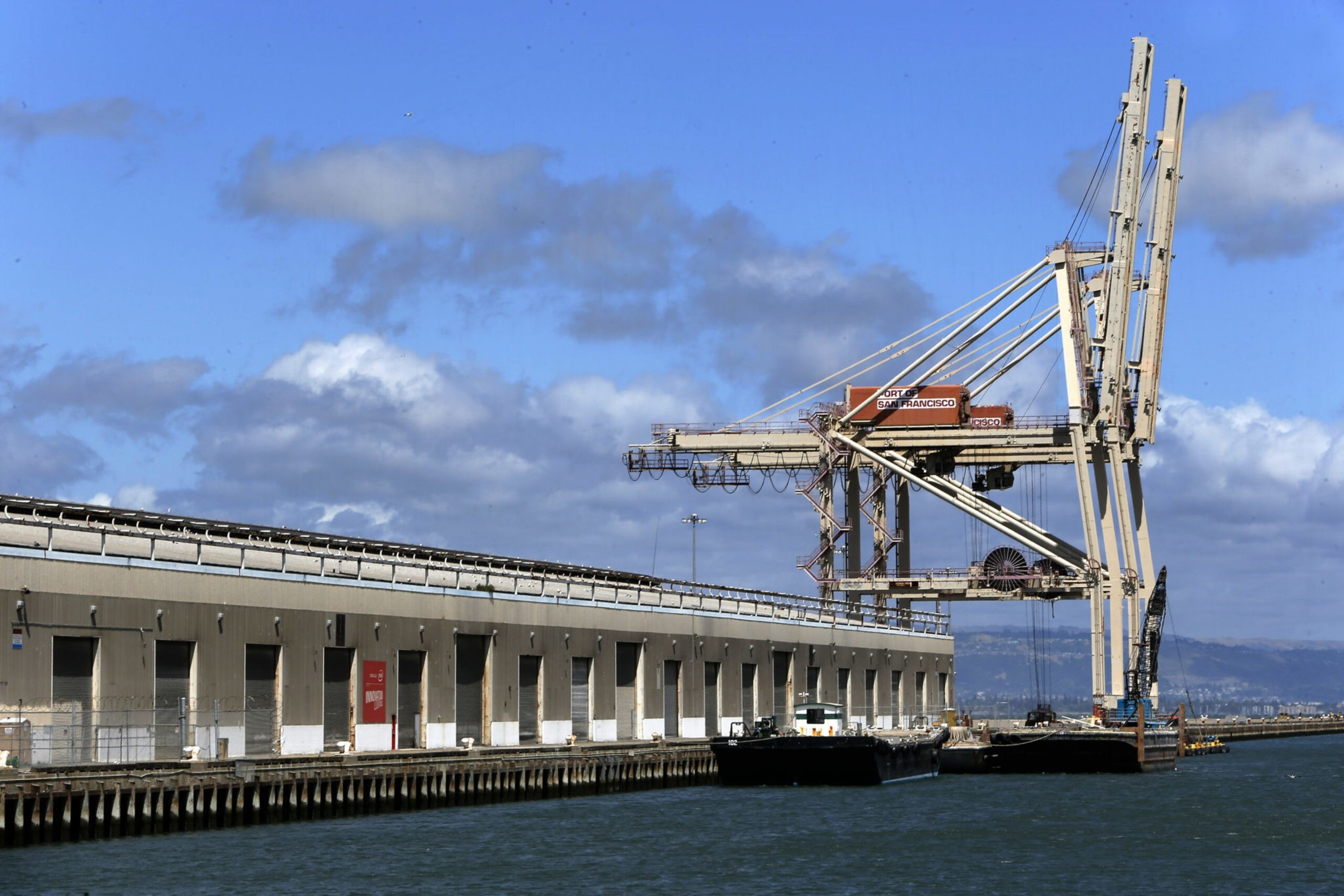 A large cargo crane at a dock extends over the water next to a long warehouse building under a partly cloudy blue sky.