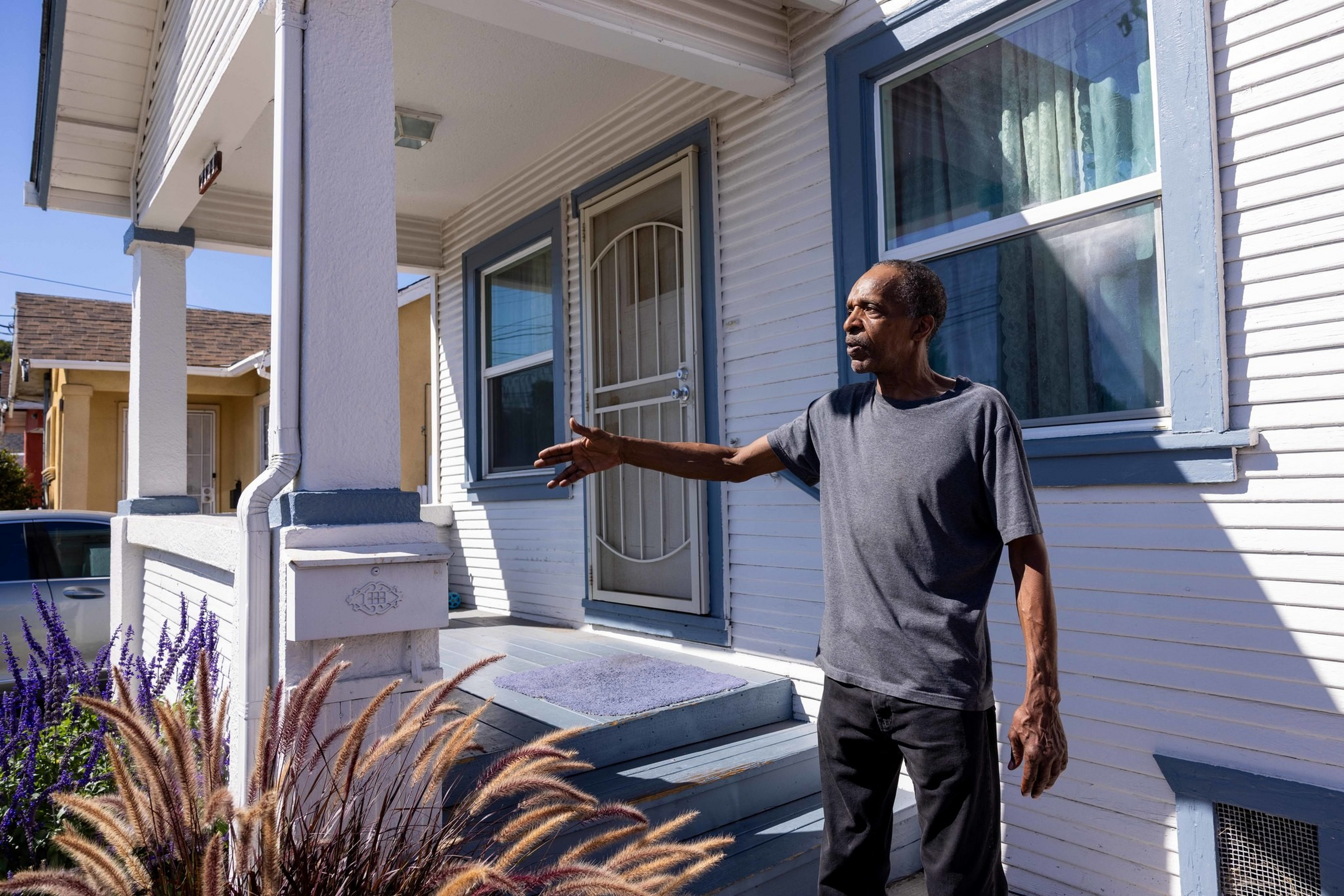 A man in a gray t-shirt and black pants stands on a porch with white siding, extending his right arm outward as if gesturing.