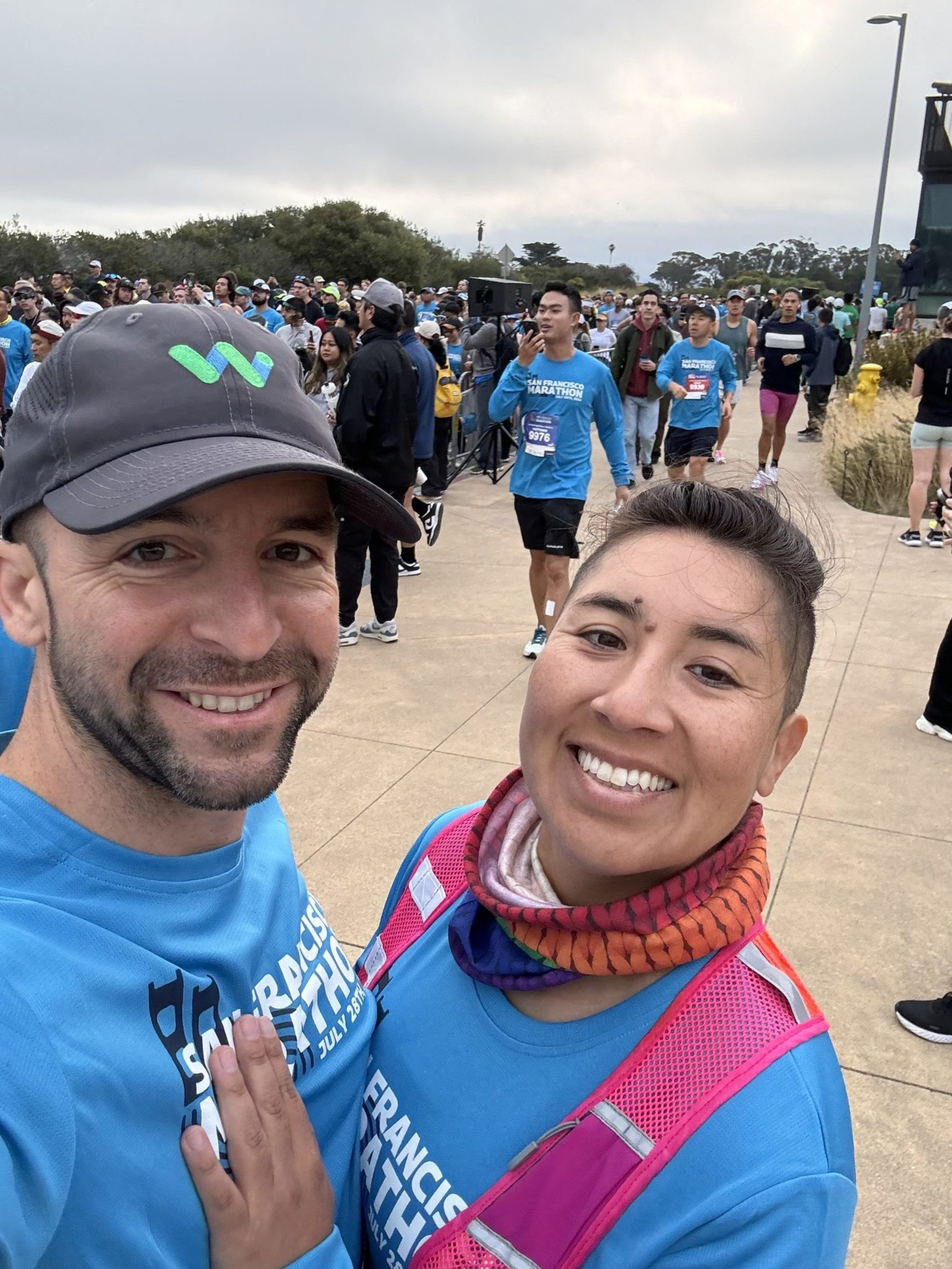 A smiling couple in matching blue marathon shirts poses for a selfie amid a large, diverse crowd of runners gathered outdoors.
