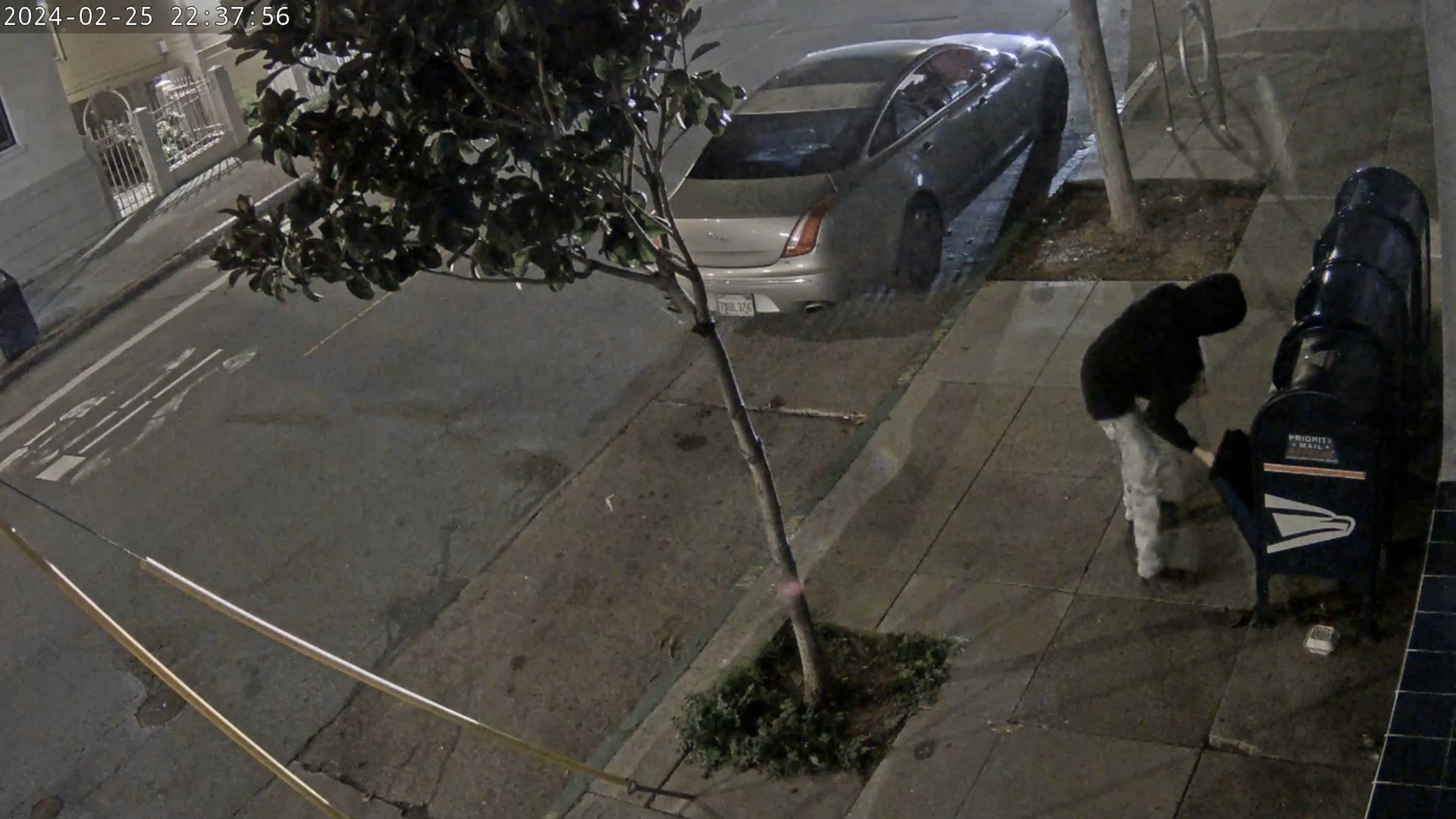 A person wearing a black hoodie is seen on the sidewalk opening blue USPS boxes