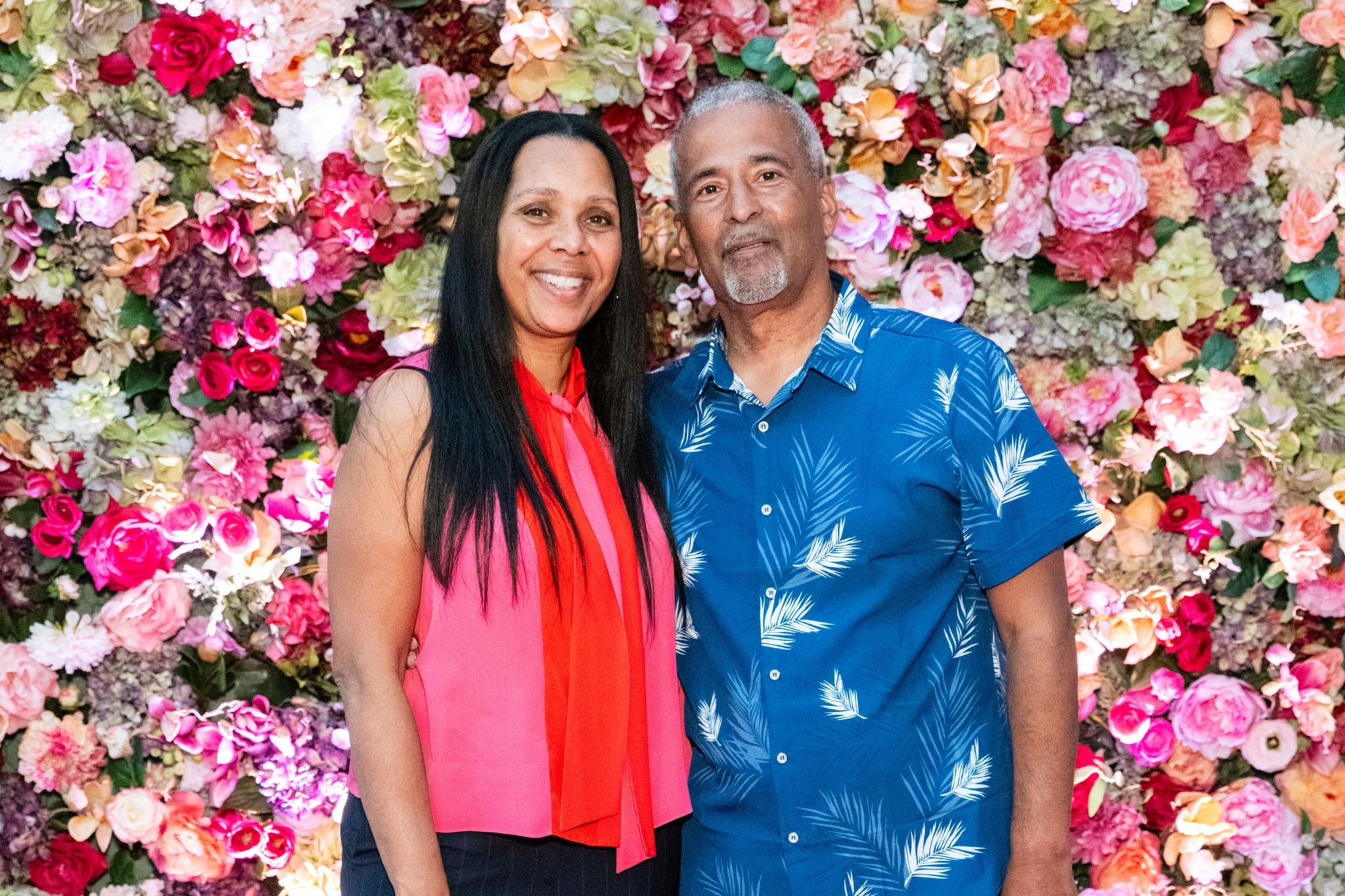 A smiling man and woman stand together against a vibrant, colorful flower wall. The woman wears a red sleeveless top, and the man wears a blue shirt with white leaf prints.