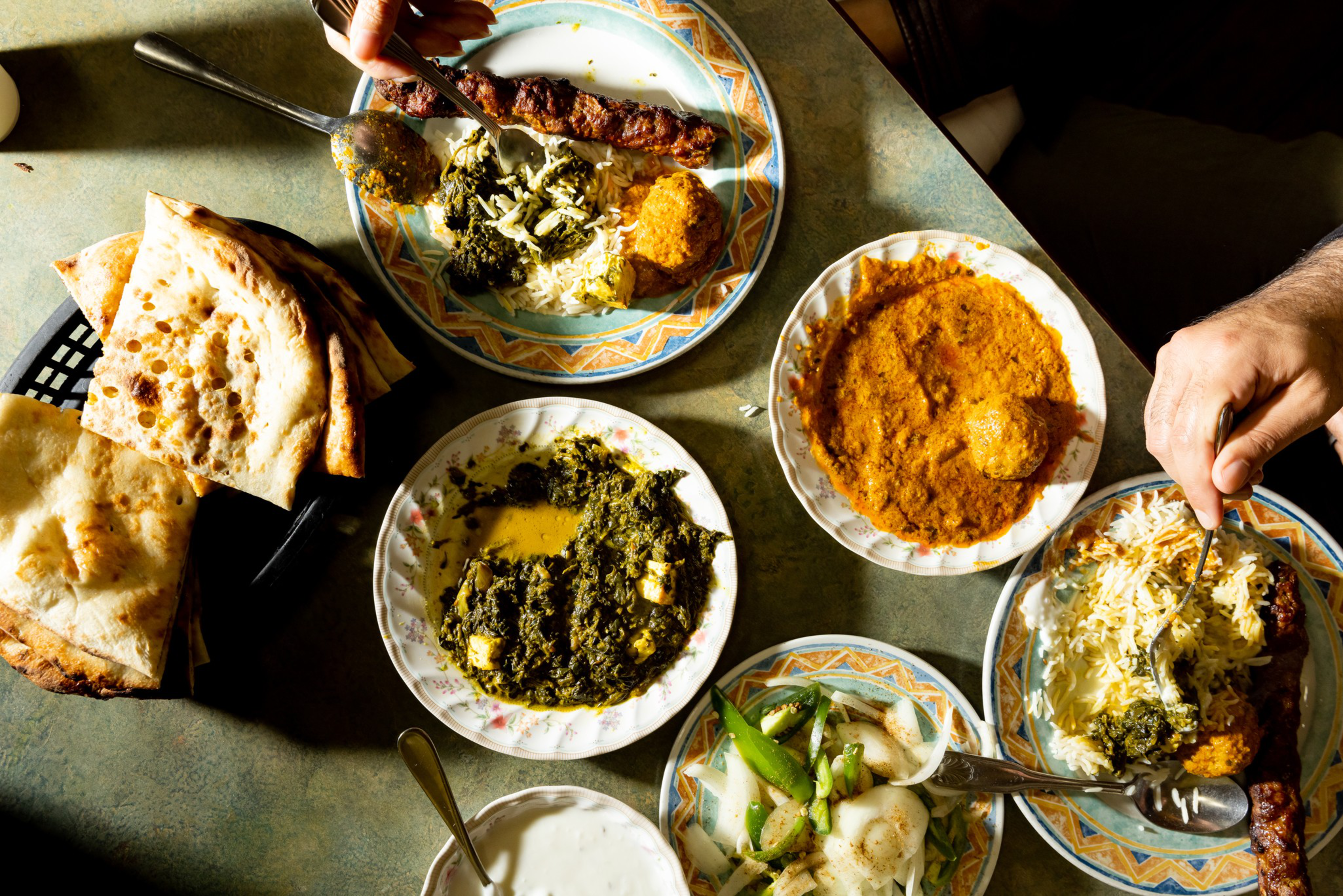 Grilled kebabs, naan bread, spinach curry with paneer, red lentil curry, rice, yogurt, and sautéed onions and peppers on a table with two people serving food.