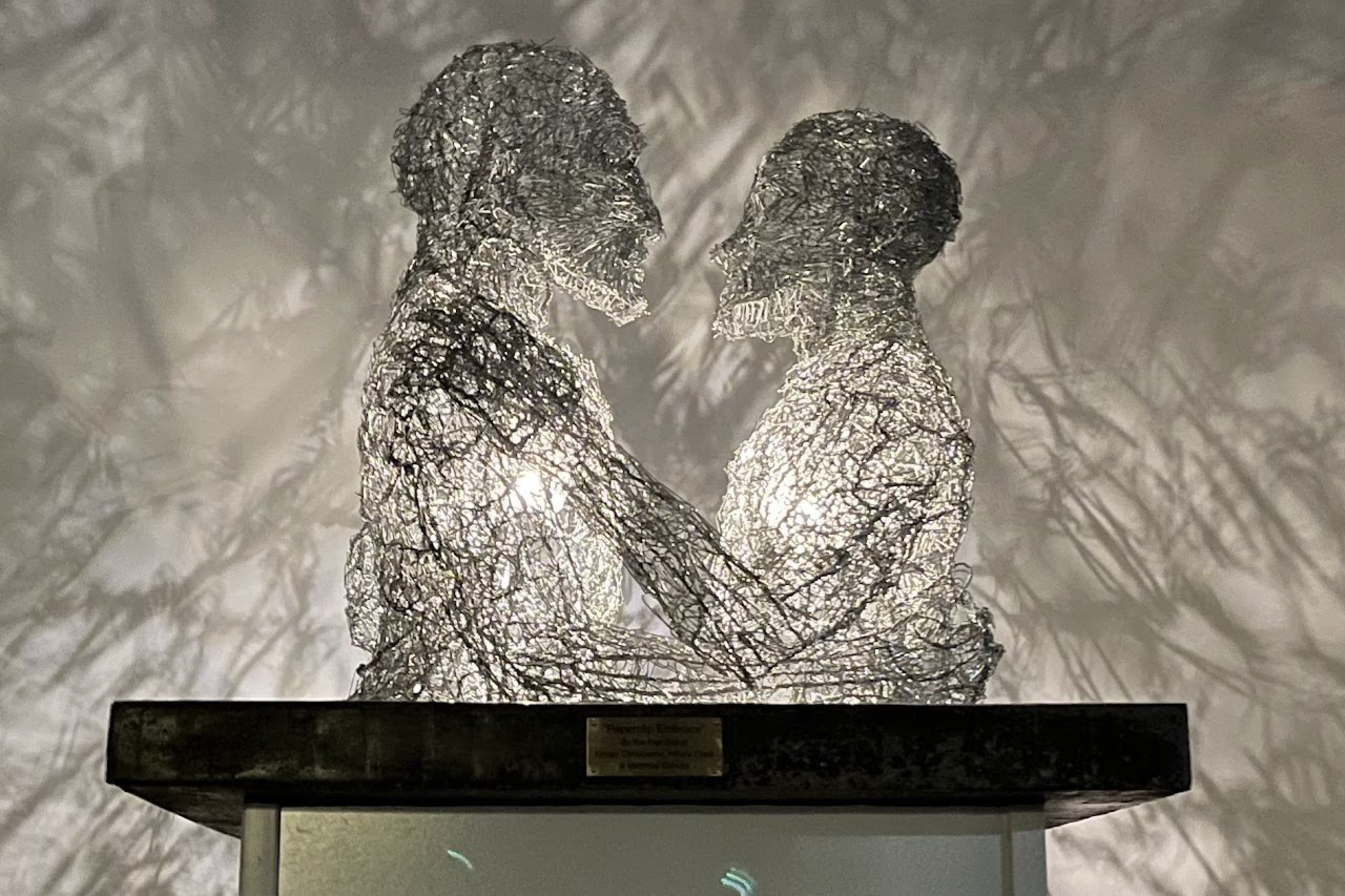 Two human figures made from intertwined wire face each other closely, their arms gently touching, with light shining through them casting shadows on the wall.