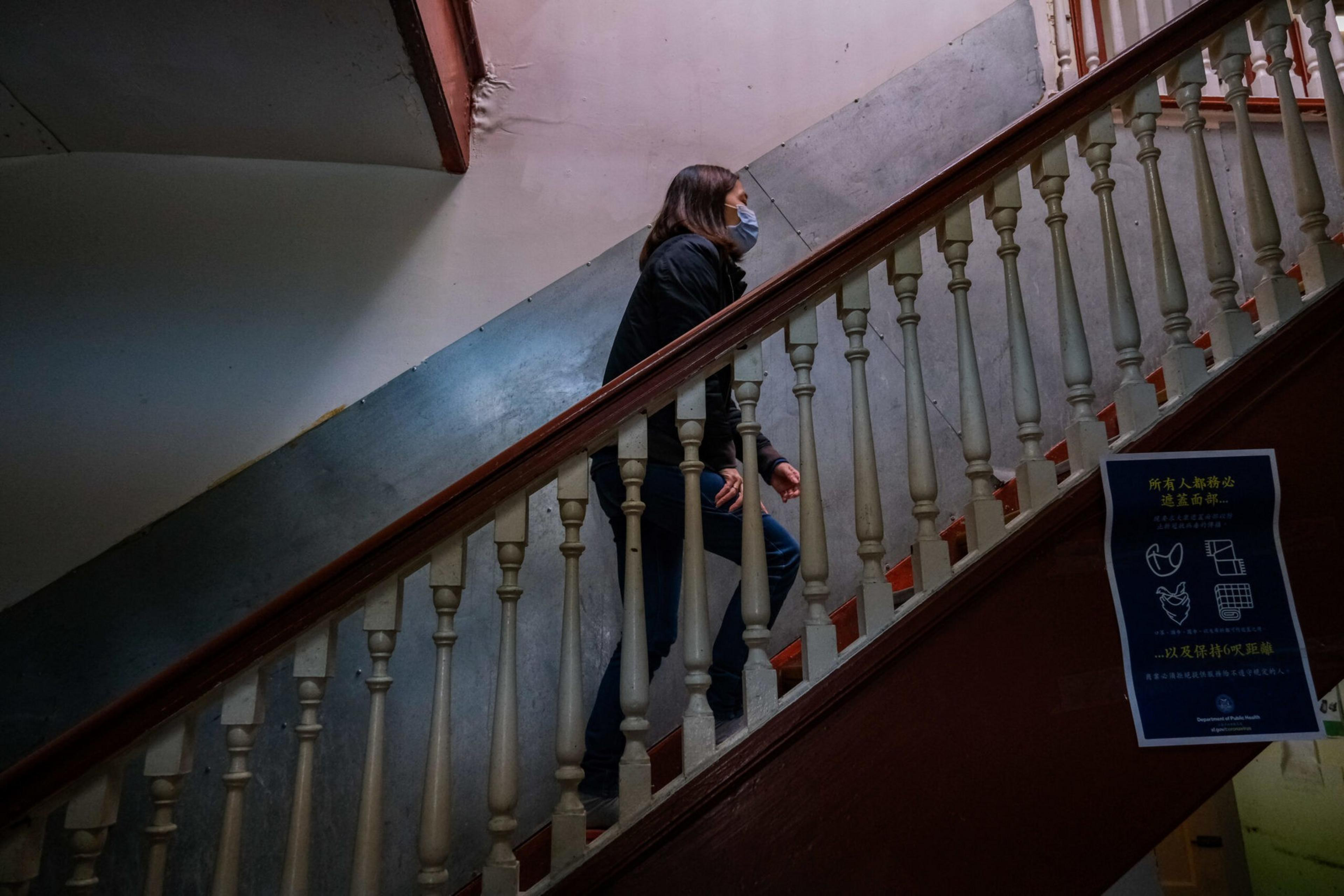A person wearing a face mask and dark clothes climbs an old wooden staircase inside a dimly lit building. A safety sign is posted on the railing.