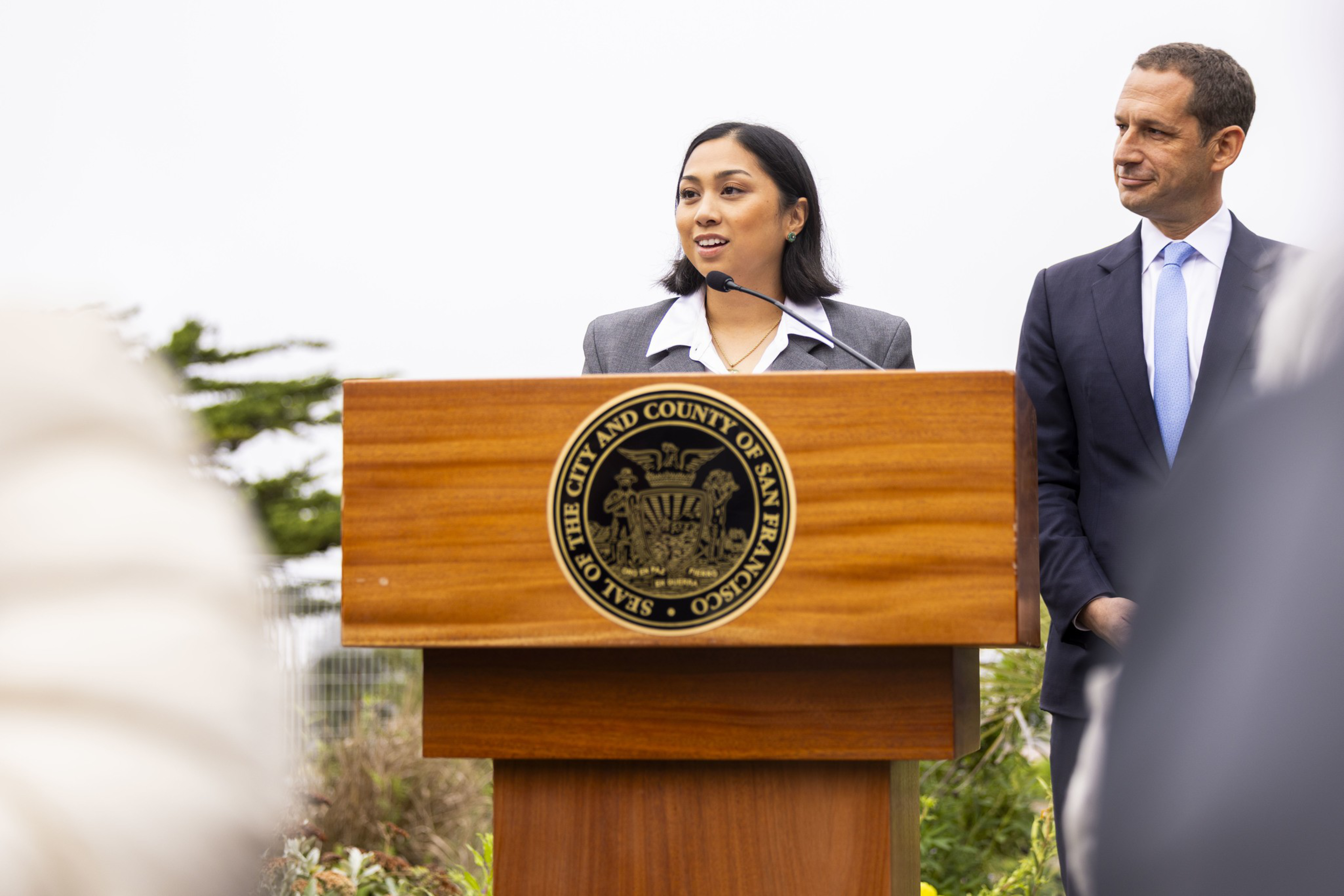 A woman in a gray suit speaks at a podium with the Seal of the City and County of San Francisco, while a man in a dark suit listens beside her.