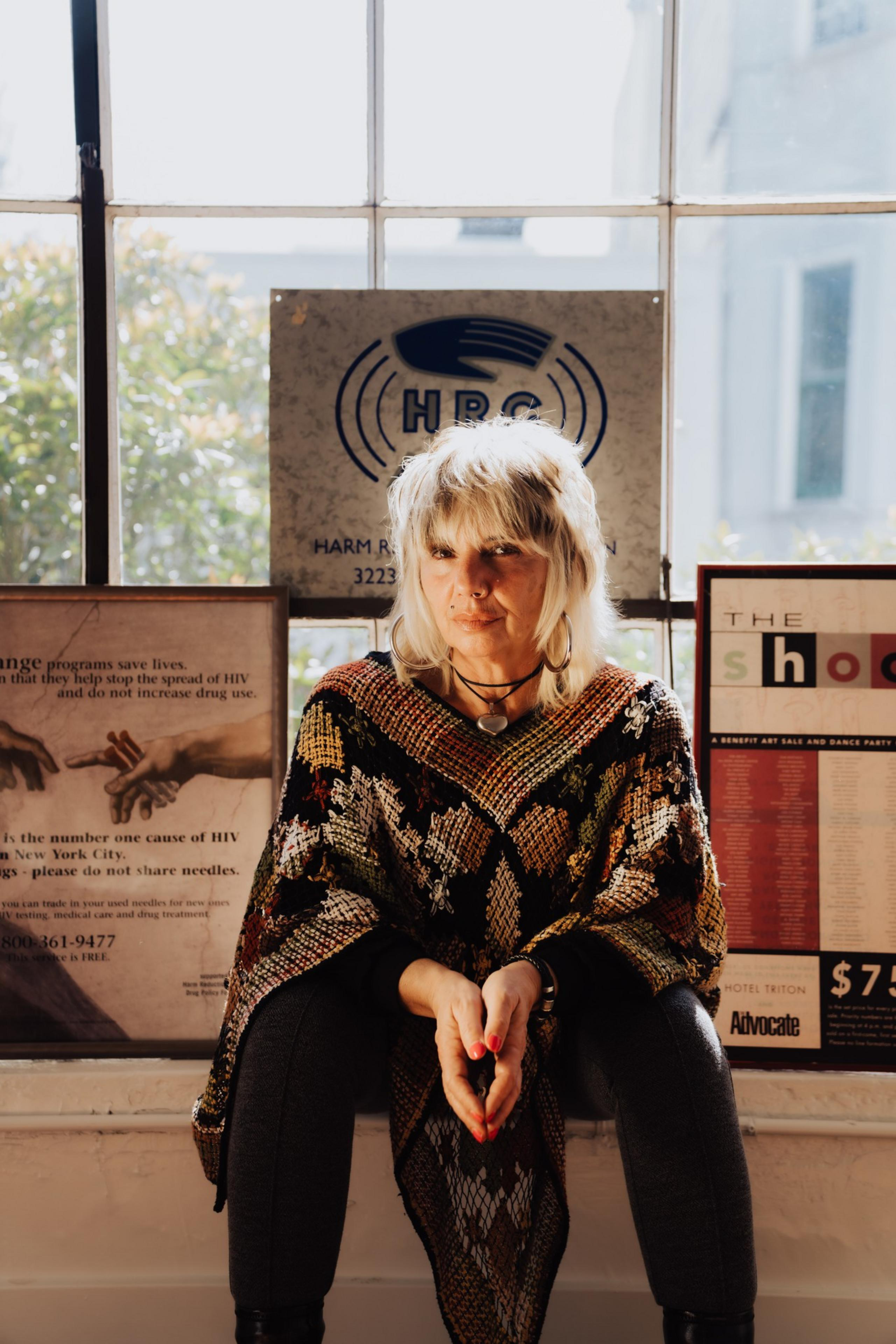 A person with light hair sits by a window, wearing a colorful patterned poncho. Behind are posters with text and logos. Light filters in, creating a warm glow.