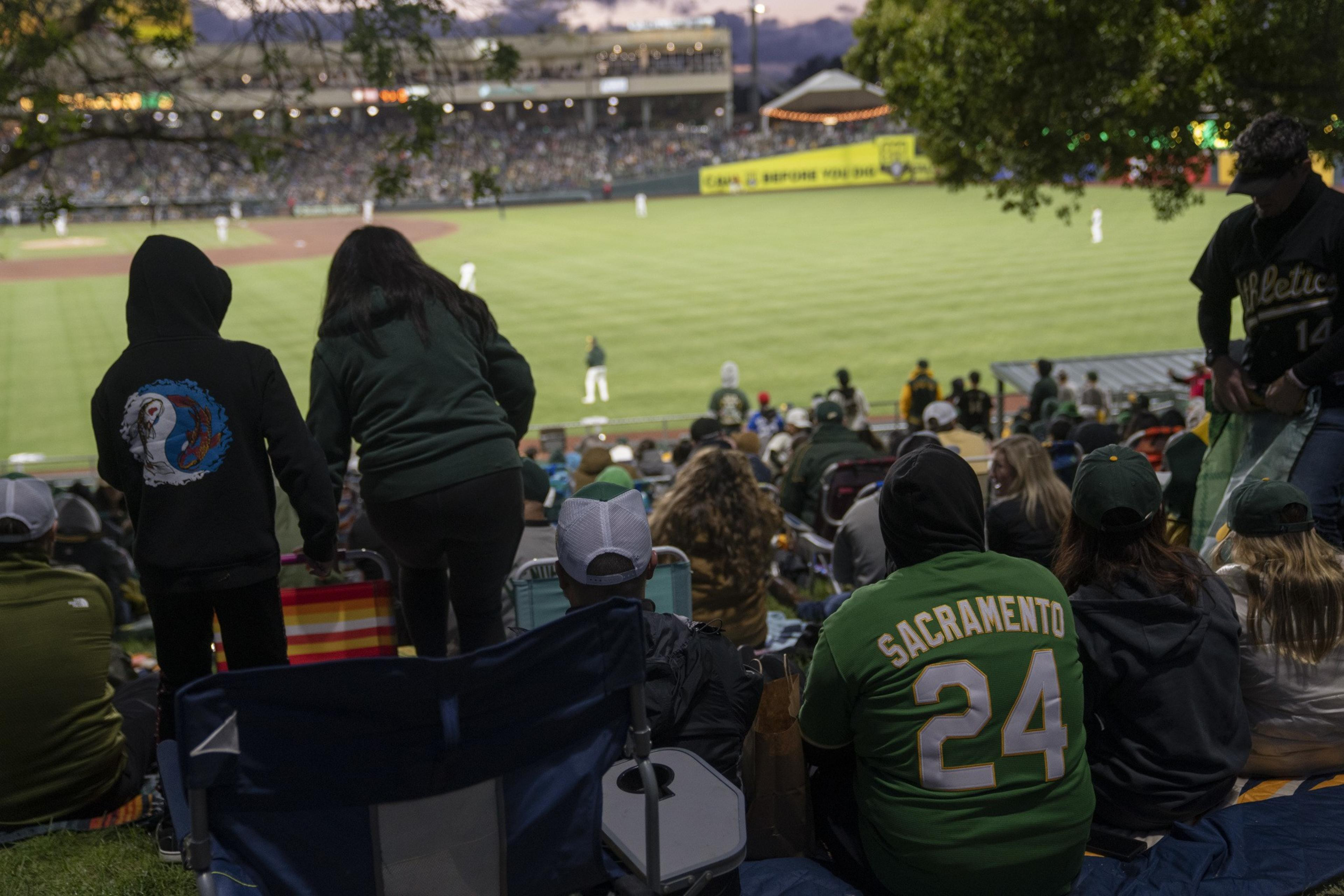 A crowd of people on a grassy hill watches a baseball game. Many wear green, including one with &quot;Sacramento 24&quot; on their jersey. The stadium is well-lit in the background.