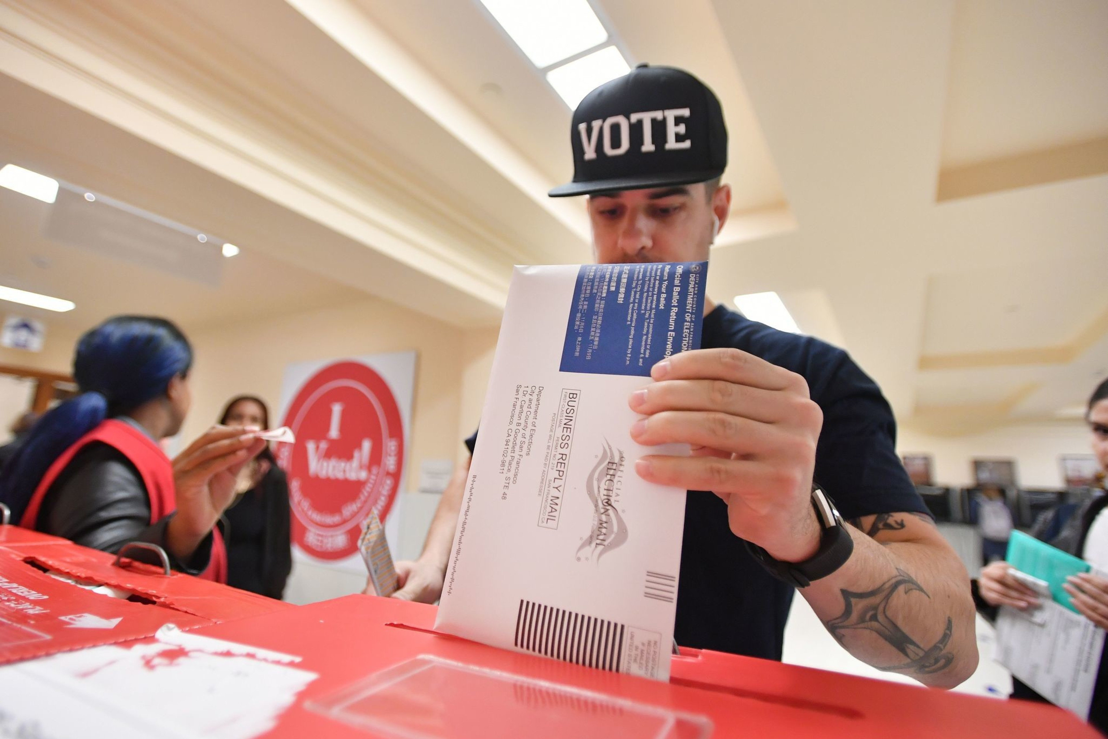 A man wearing a hat drops a ballot envelope into a container.