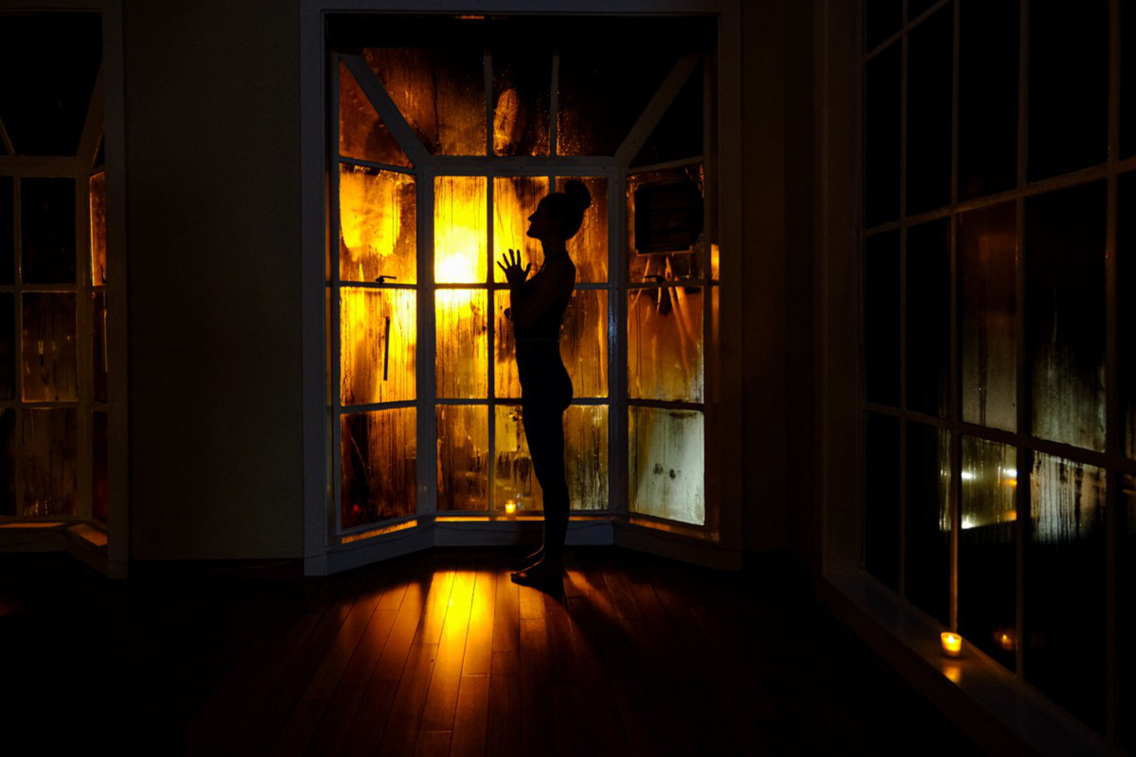 A silhouetted person stands near a foggy window glowing with warm orange light, their hands raised as if touching the glass.