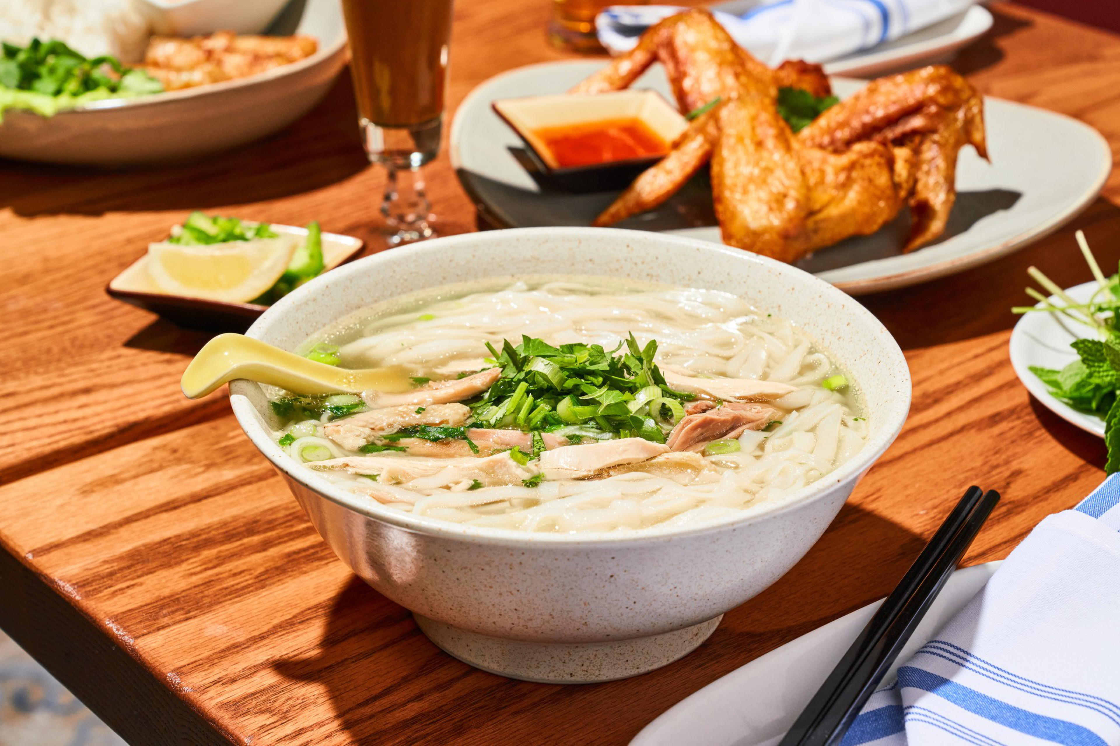 A bowl of noodle soup with herbs and meat sits on a wooden table, next to fried chicken, lemon wedges, and fresh greens, with chopsticks nearby.