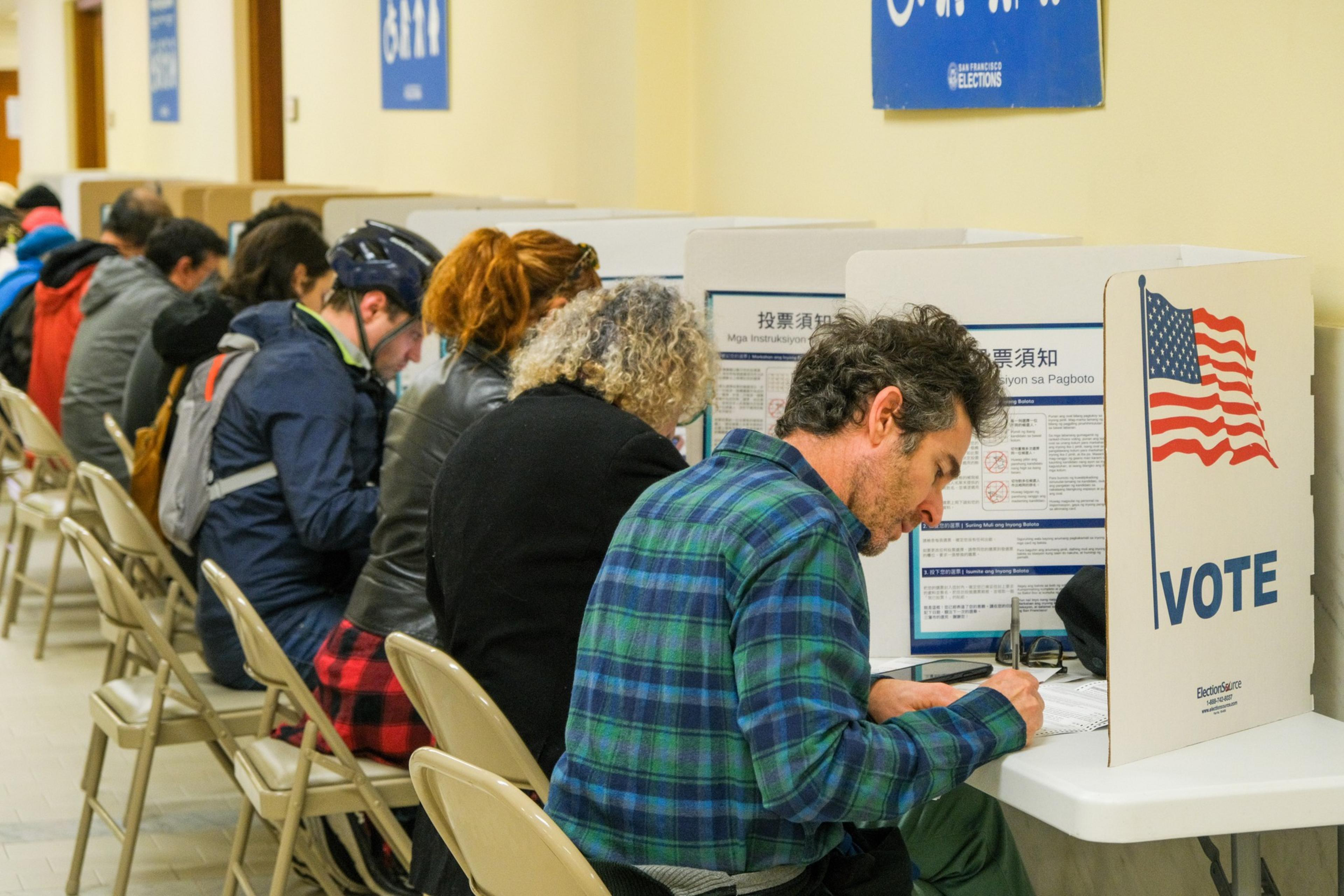 A row of people is seated in partitioned booths, casting votes. The dividers feature an American flag and "VOTE" text. Signs in multiple languages are visible above them.