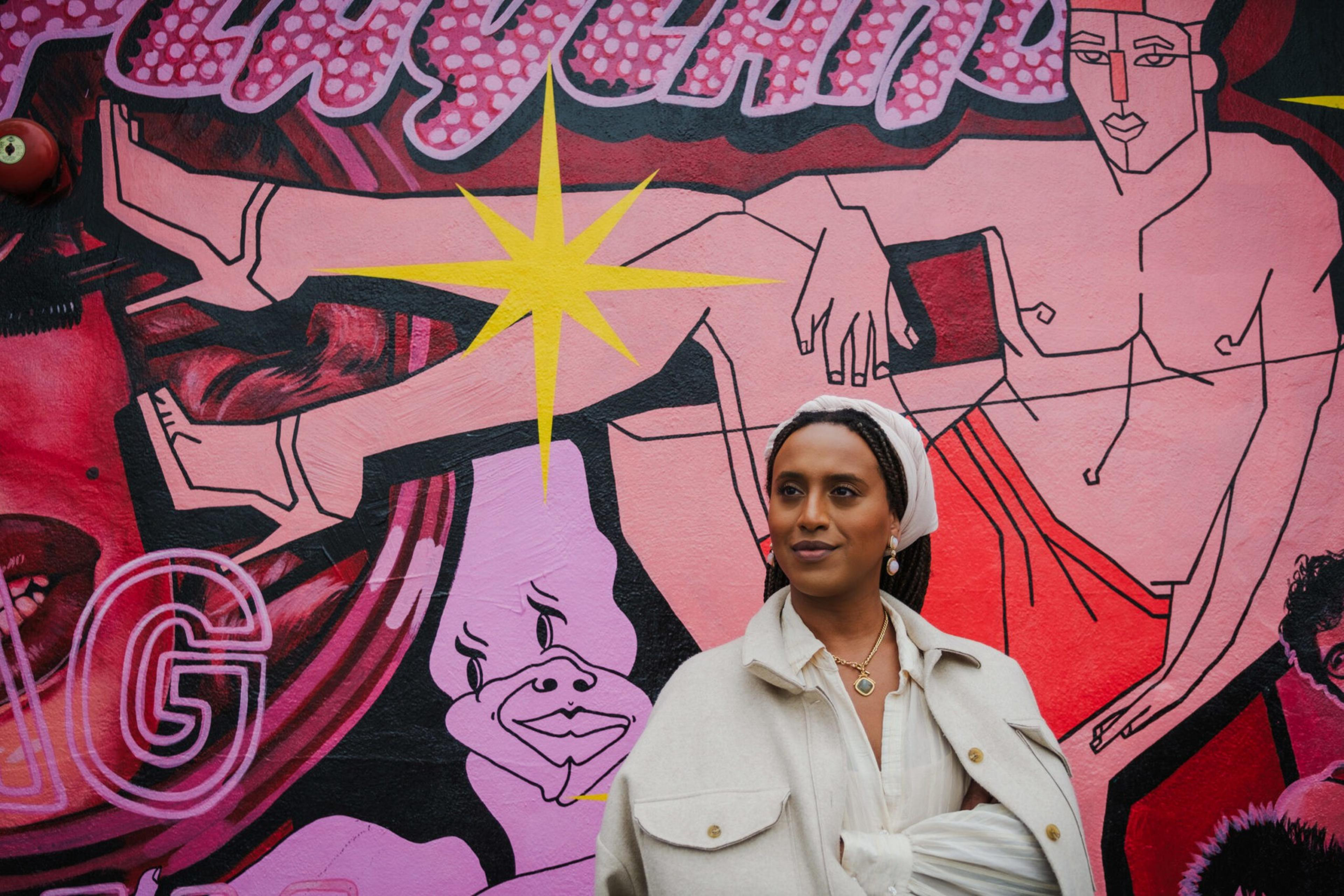 A woman in a light coat and headband stands in front of a vibrant mural featuring stylized figures, bold colors, and a bright yellow starburst.