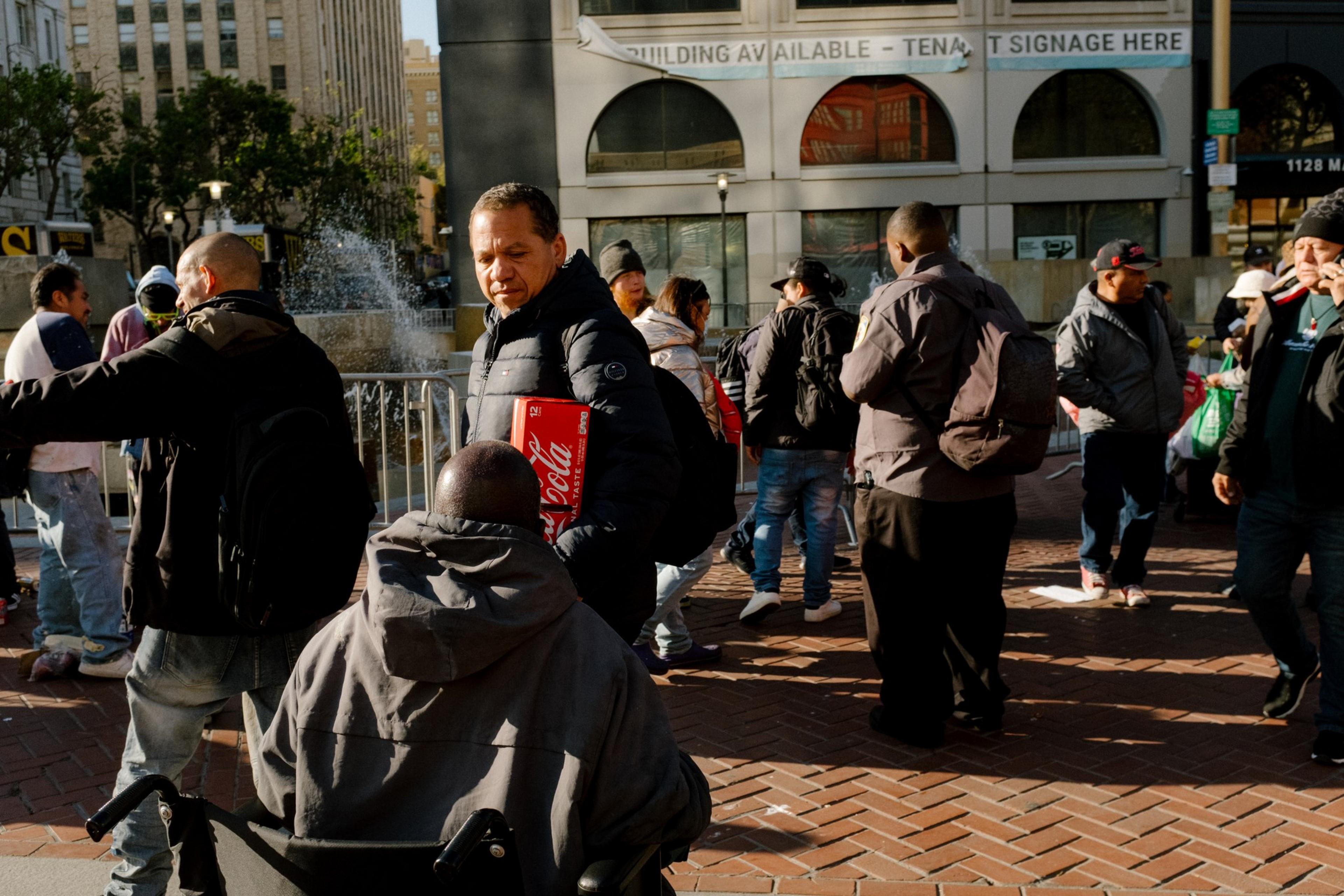 A busy street scene with people walking, one in a wheelchair, and a fountain in the background.