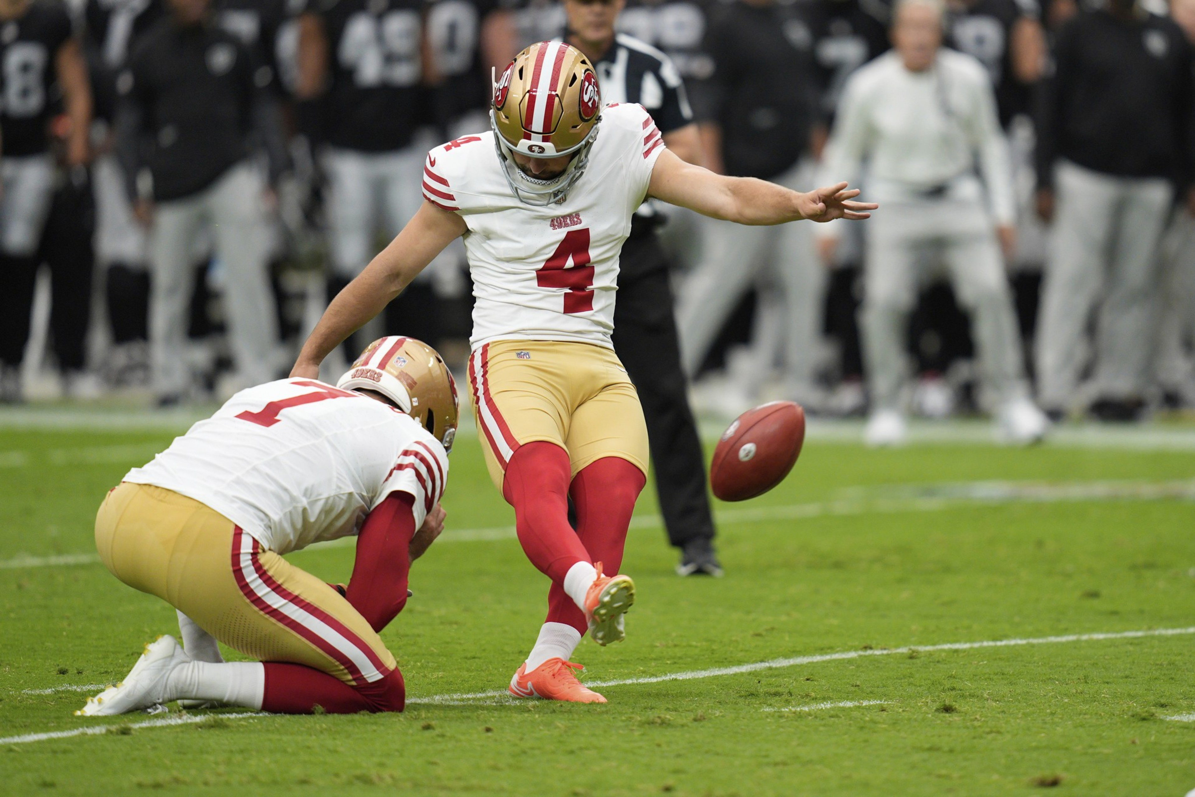 A football player wearing number 4 kicks the ball while a teammate holds it on the ground during a game.