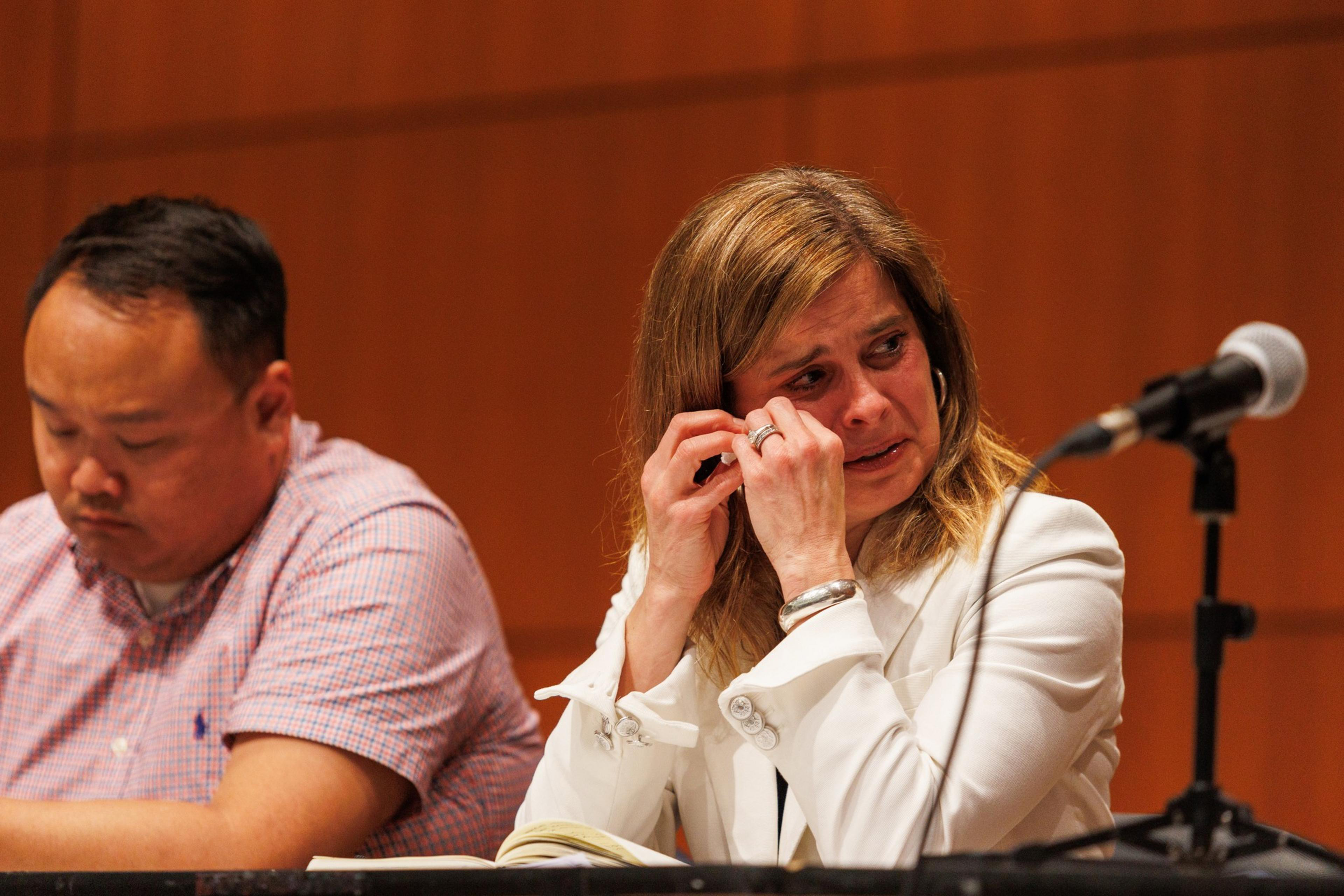 A woman in white, wiping tears away, sits beside a focused man at a panel with microphones.