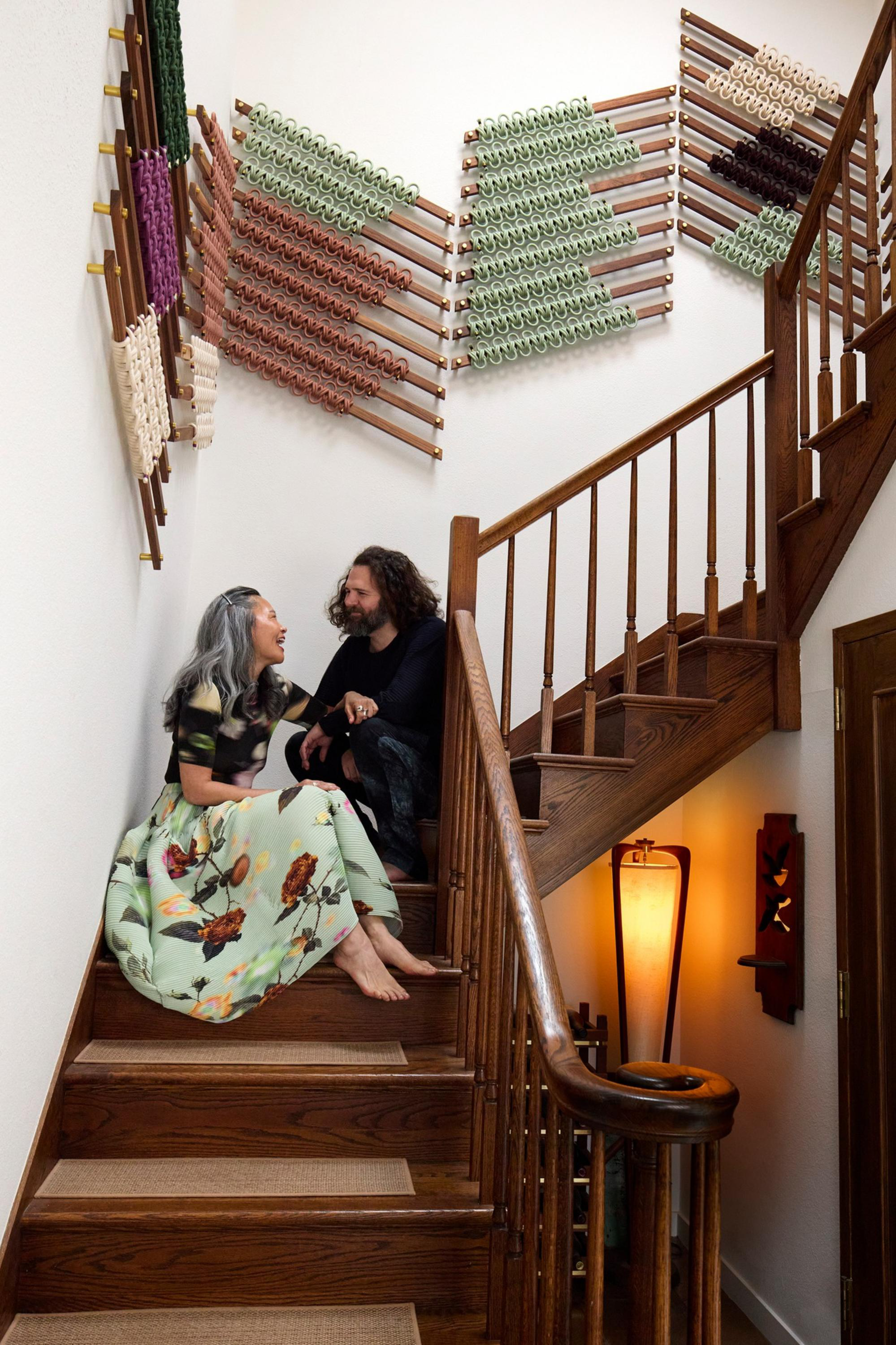 Two people sit barefoot on wooden stairs, smiling and talking, with colorful woven wall art above and a warm glowing lamp below.