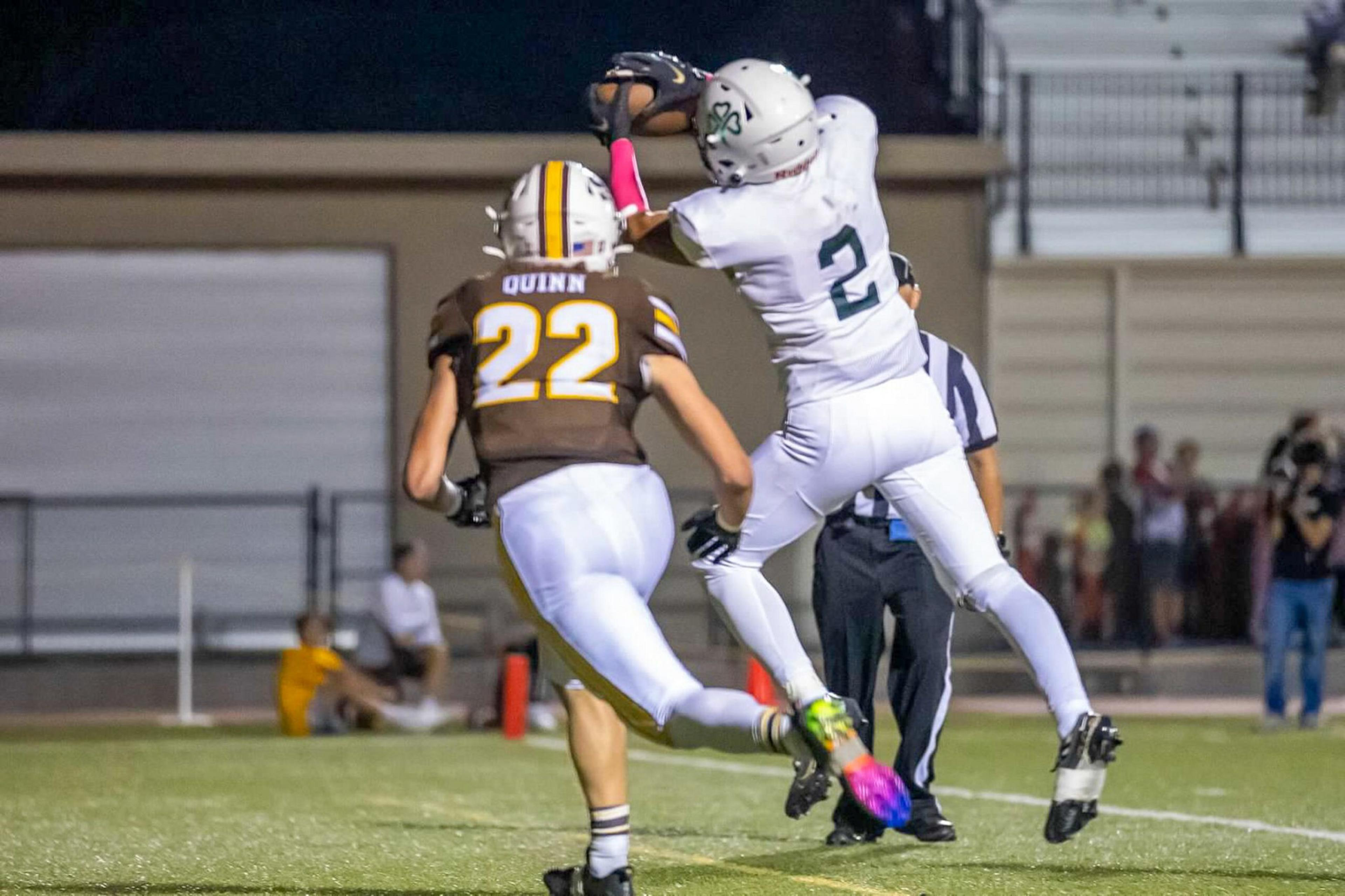 A football player in white jersey number 2 leaps to catch the ball while a player in brown jersey number 22 runs closely behind on a field.