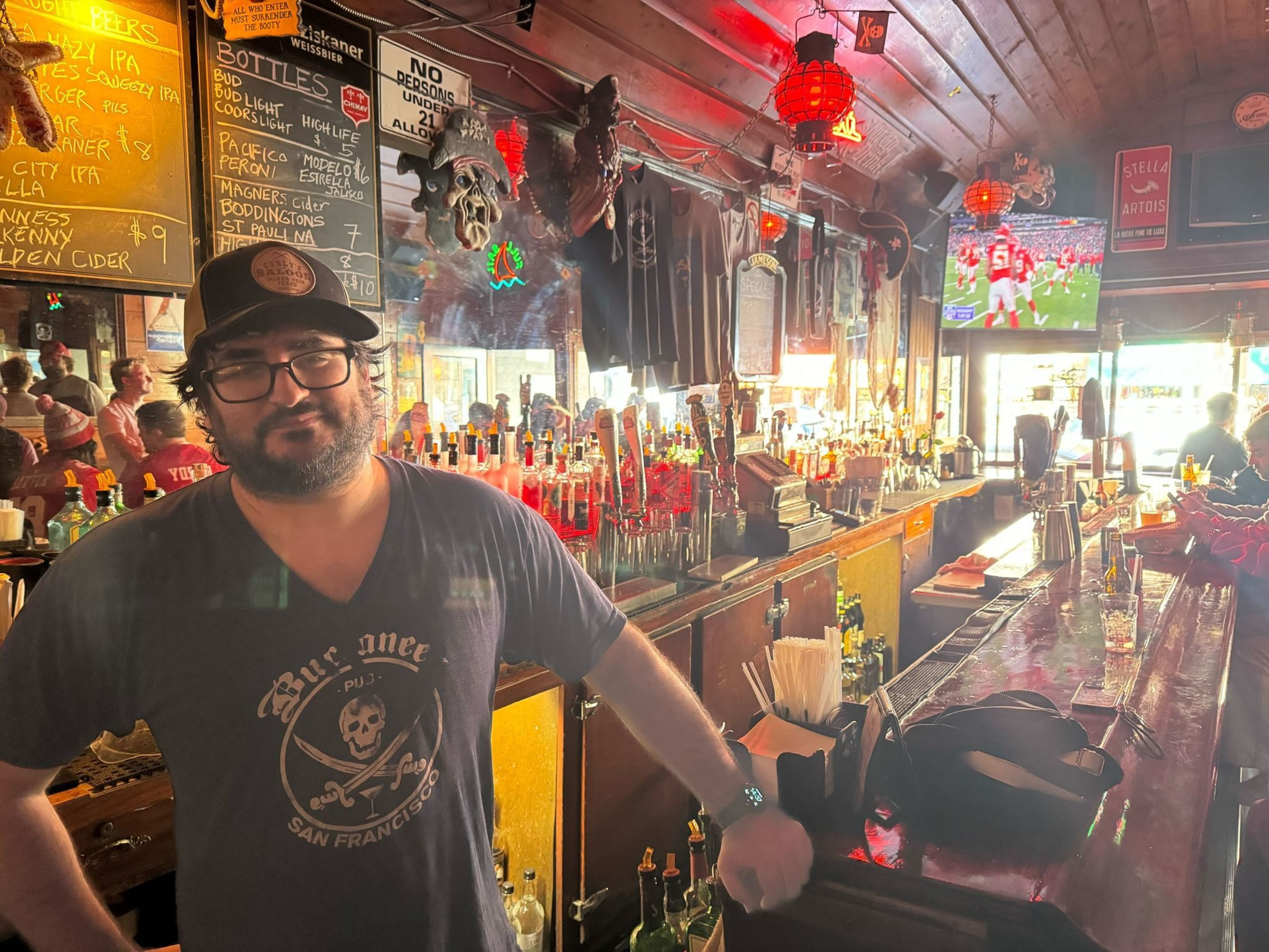 A man stands at a bar lit by red lanterns, with TVs showing sports and a vibrant atmosphere.