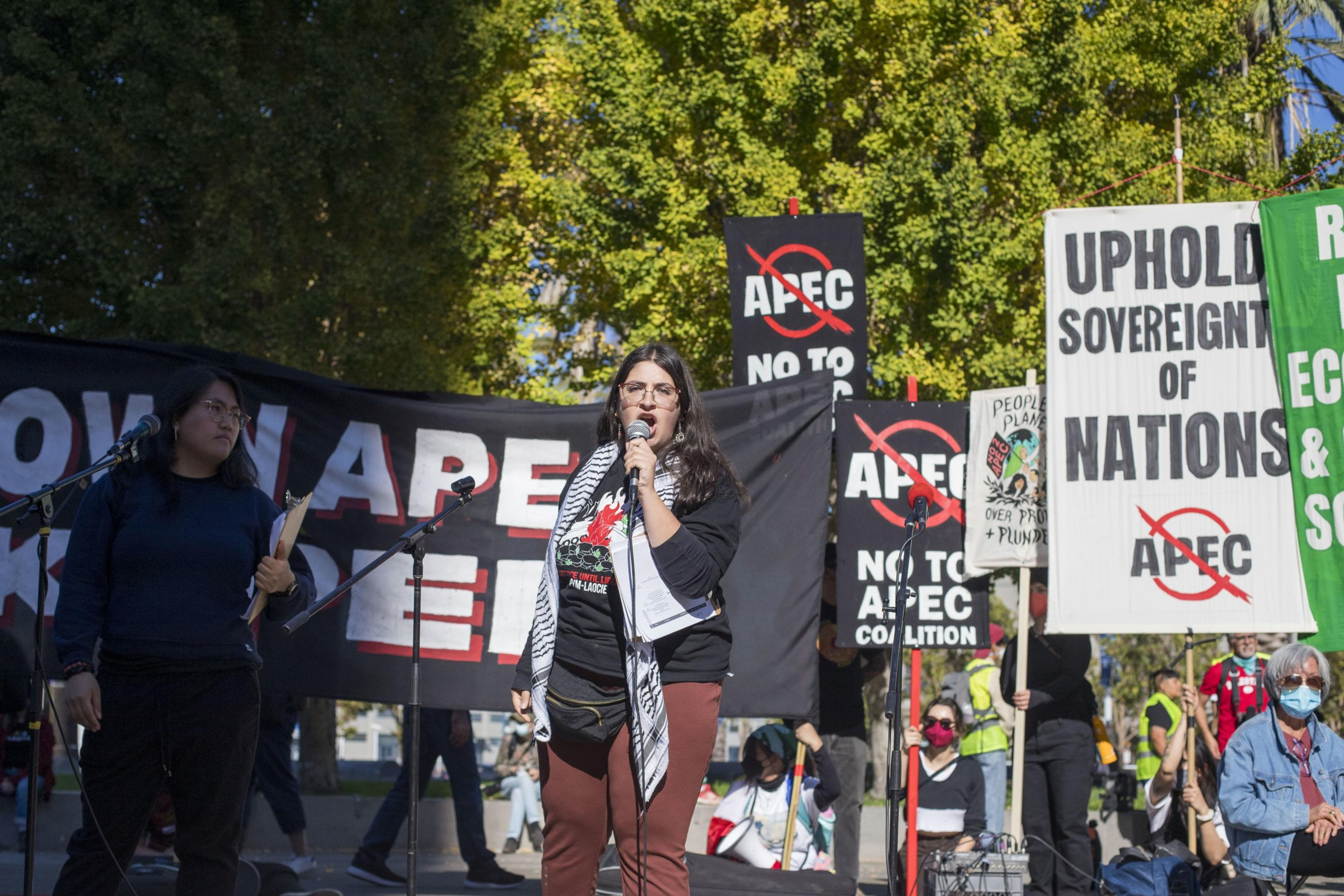 A person is giving a speech on a microphone in front of signs protesting the APEC summit.