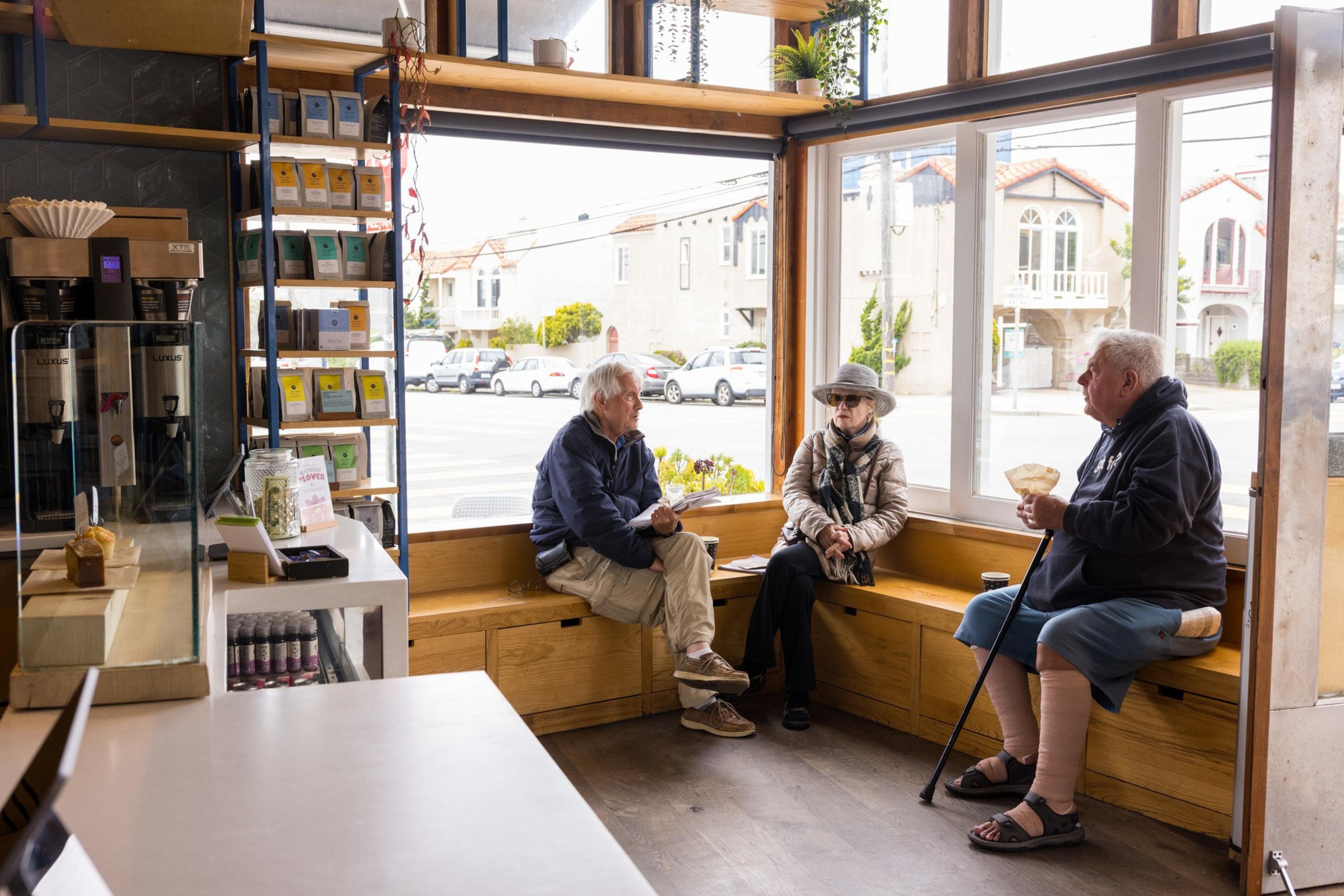 Three people sit in a cozy café by a large window. Coffee equipment and shelves with colorful bags are visible. Outside, cars line the street.