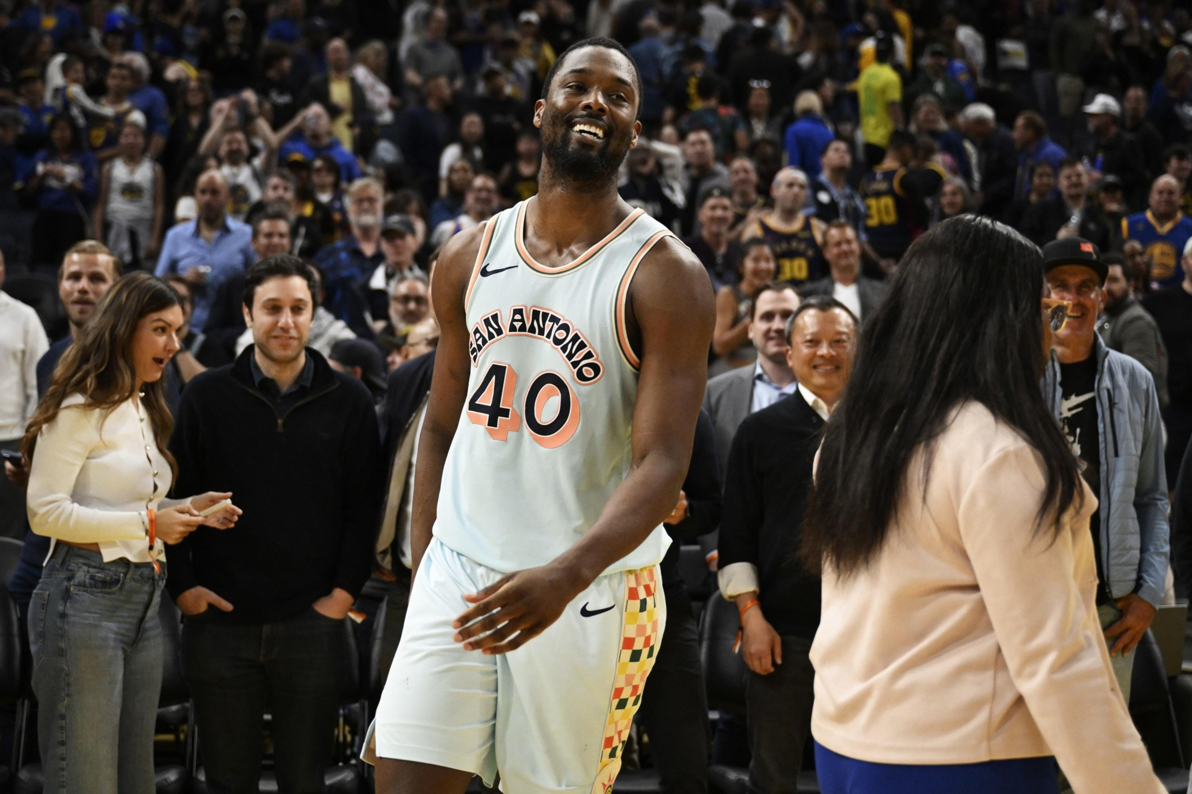 A basketball player wearing a "San Antonio" jersey smiles amid a cheering crowd. People around are excited, and the atmosphere is lively and celebratory.