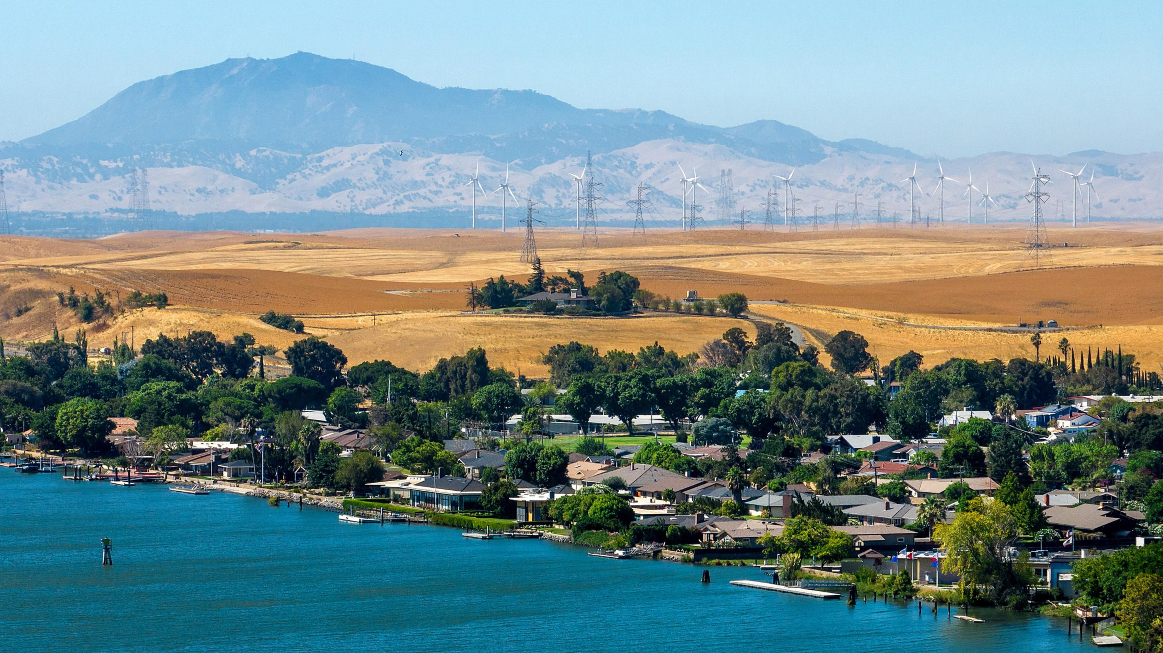 The image shows a scenic view of a neighborhood by a lake, with a backdrop of lush greenery, golden fields, wind turbines, and mountains under a clear blue sky.