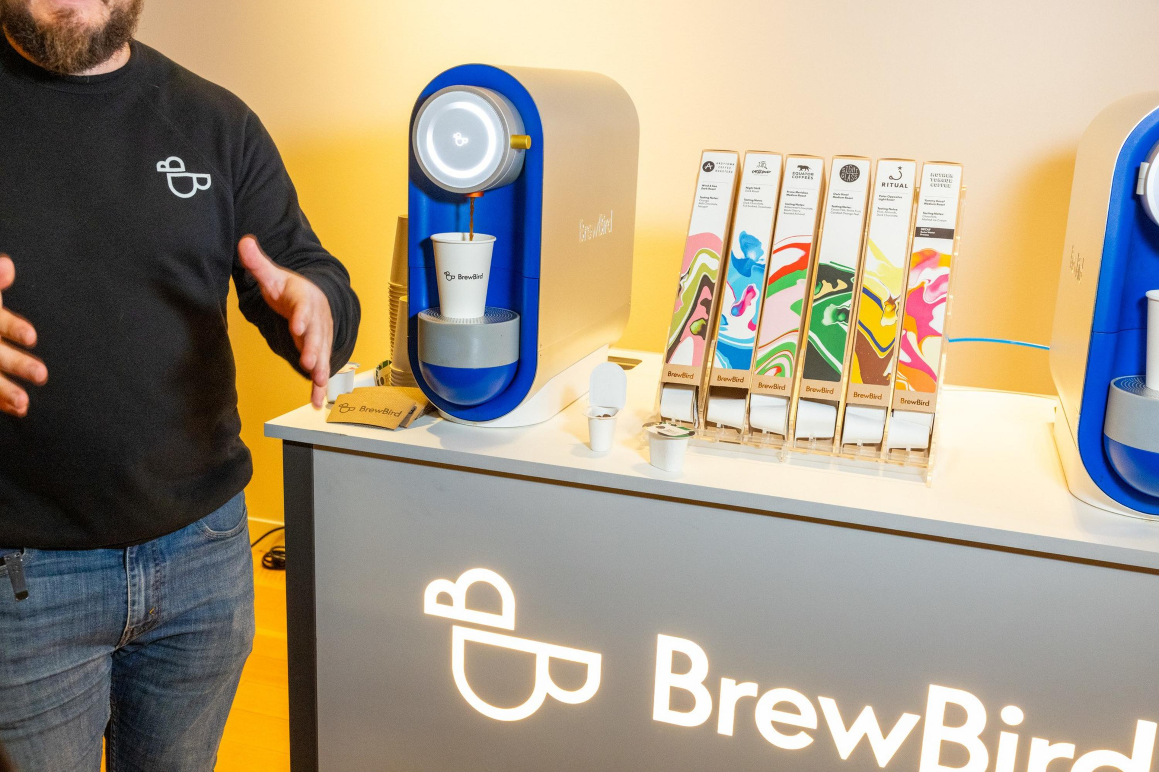 A person stands next to a BrewBird coffee machine pouring into a cup, with six coffee pods in colorful packaging displayed on a counter.
