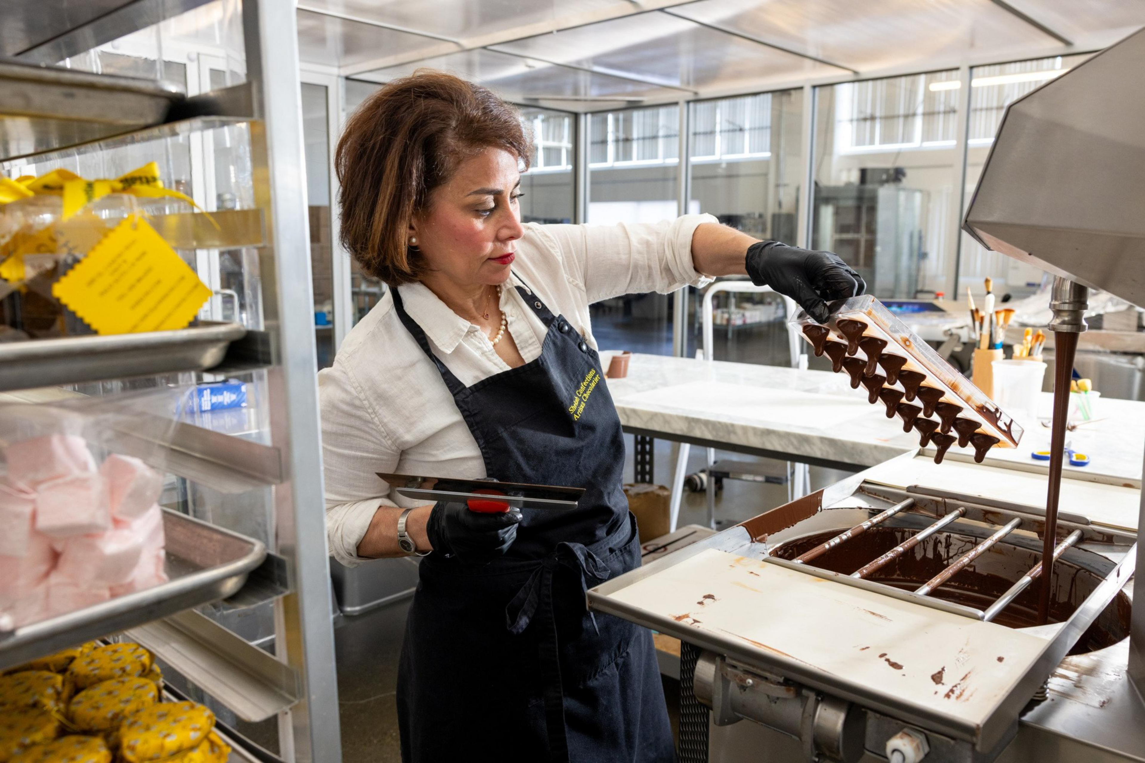 A woman in a white shirt and black apron carefully fills chocolate molds from a machine while standing in a commercial kitchen.