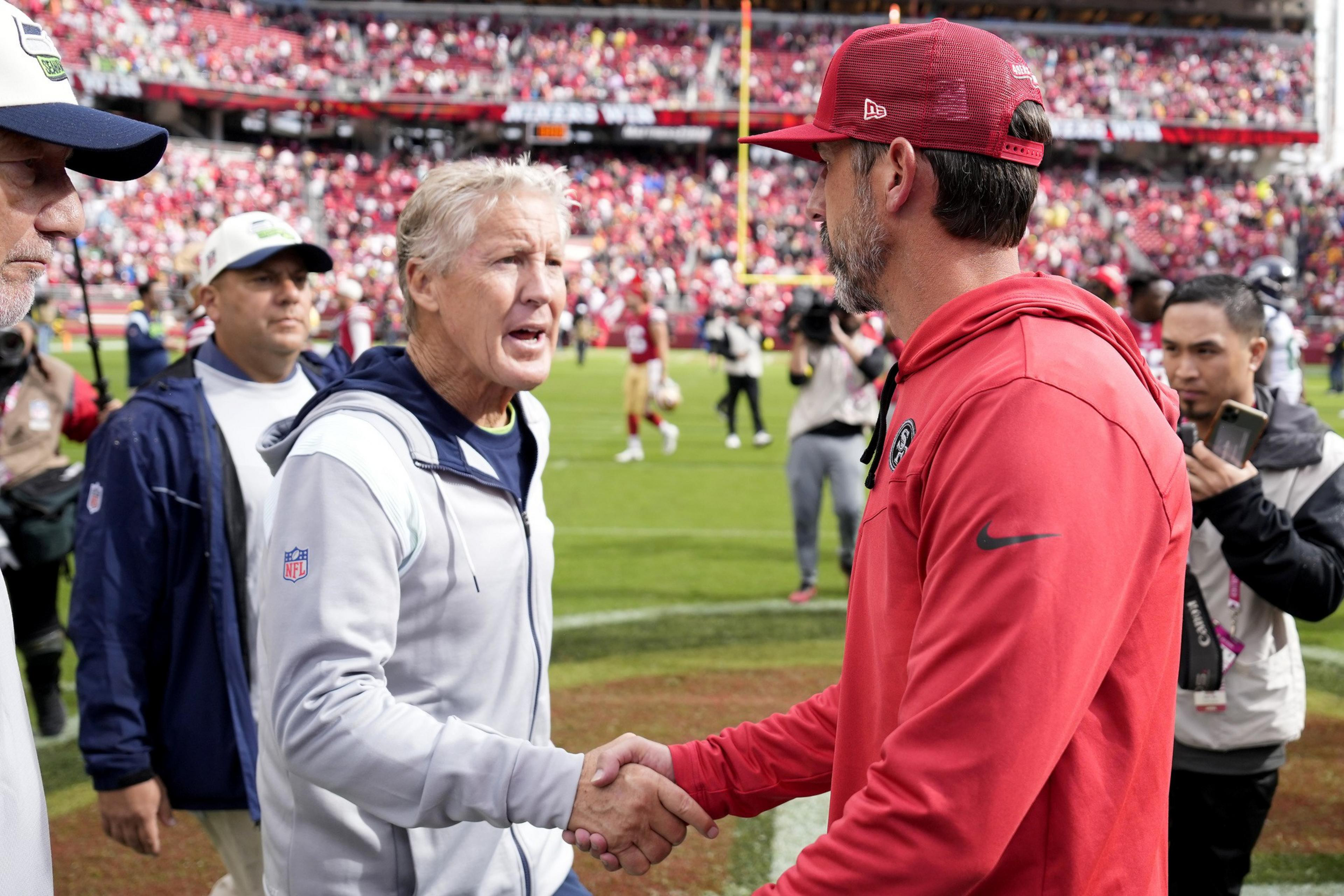 Two men, one in a light jacket and the other in a red hoodie and cap, shake hands on a football field, surrounded by people and a stadium crowd.
