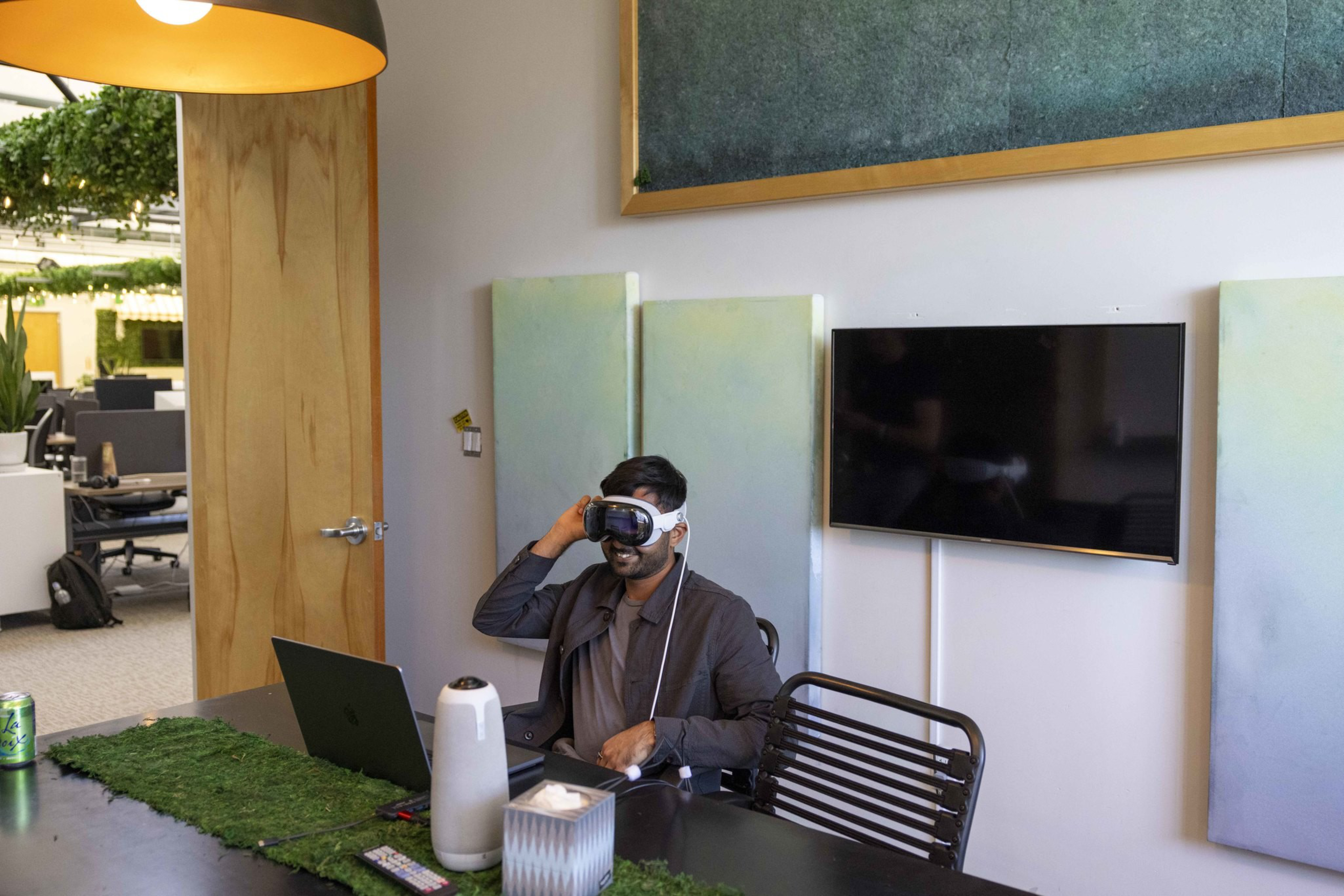 A person wearing VR goggles sits at a desk with a laptop in a modern, plant-decorated office, featuring a large TV and soft green color panels on the wall.