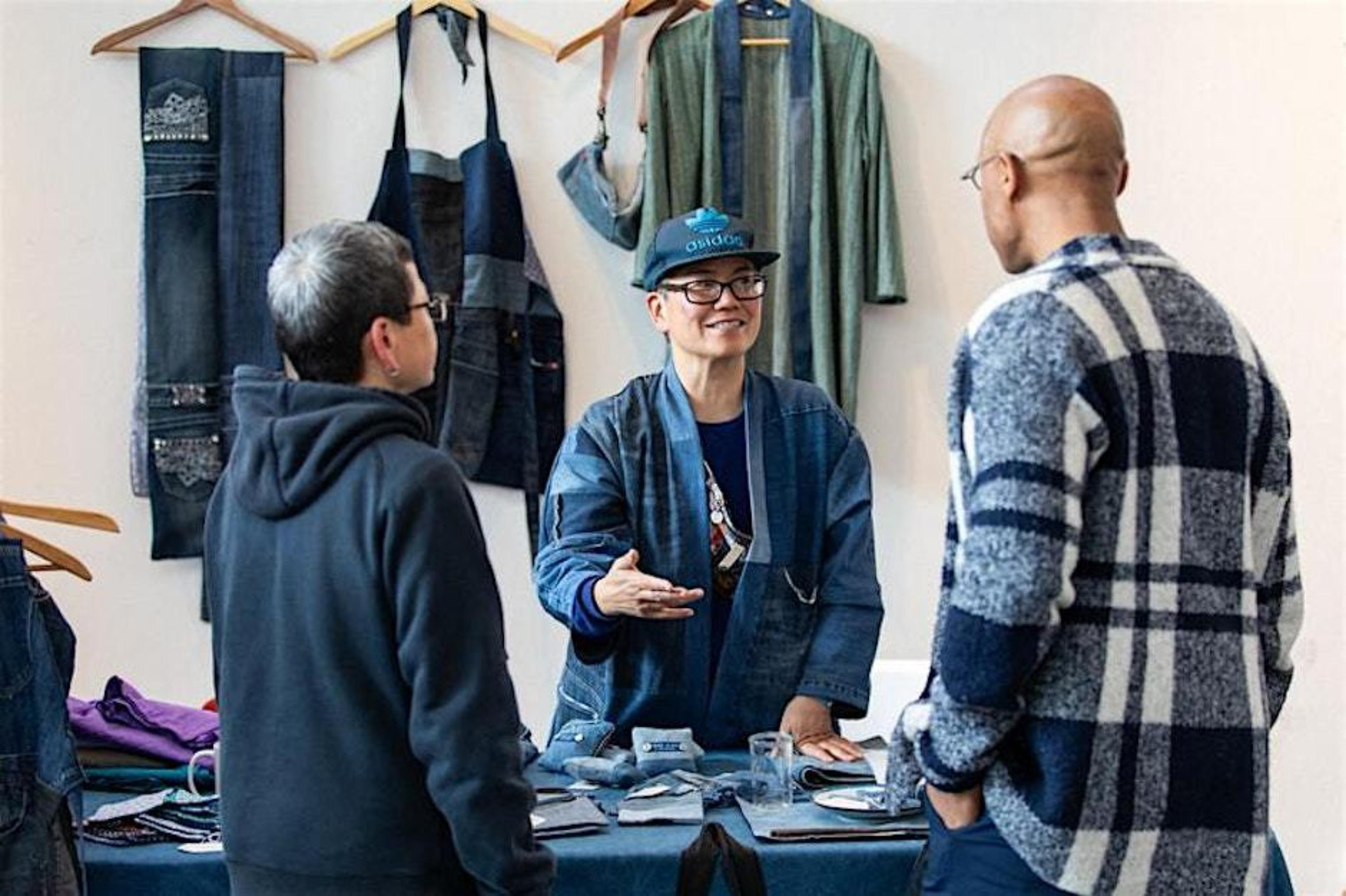 Three people are discussing in front of a table with fabric items. One wears a cap and gestures while speaking. Garments hang on the wall behind them.