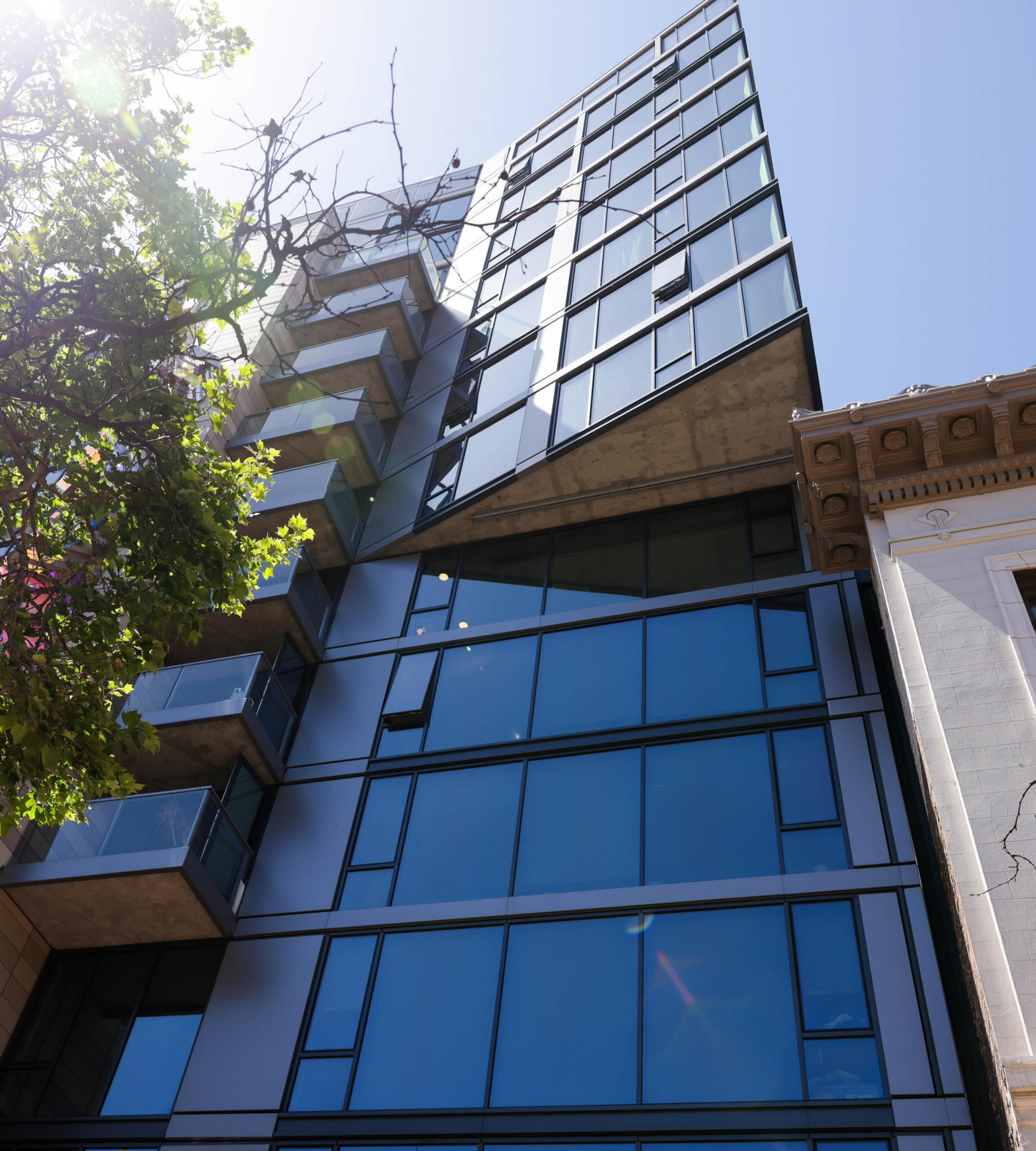 Modern glass building with reflective windows beside a traditional building, viewed from below with a tree and clear sky.