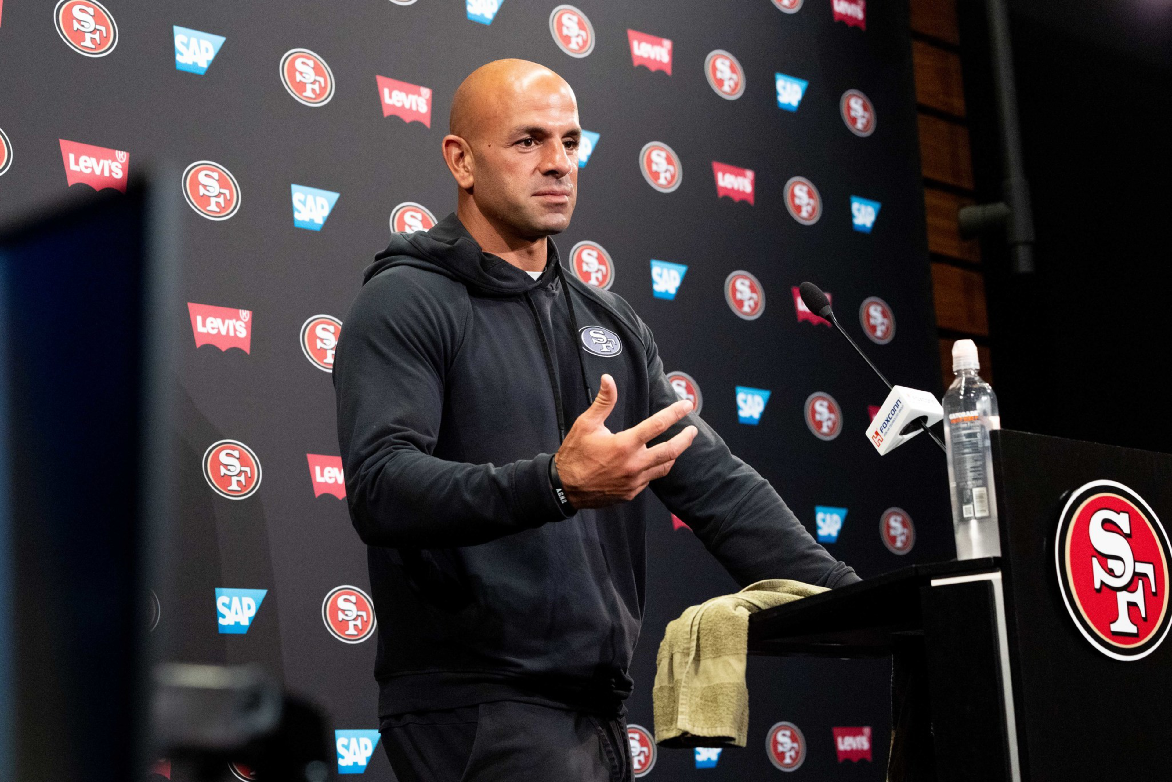 A bald man in a black San Francisco 49ers hoodie speaks at a press conference podium with a 49ers logo and water bottle.