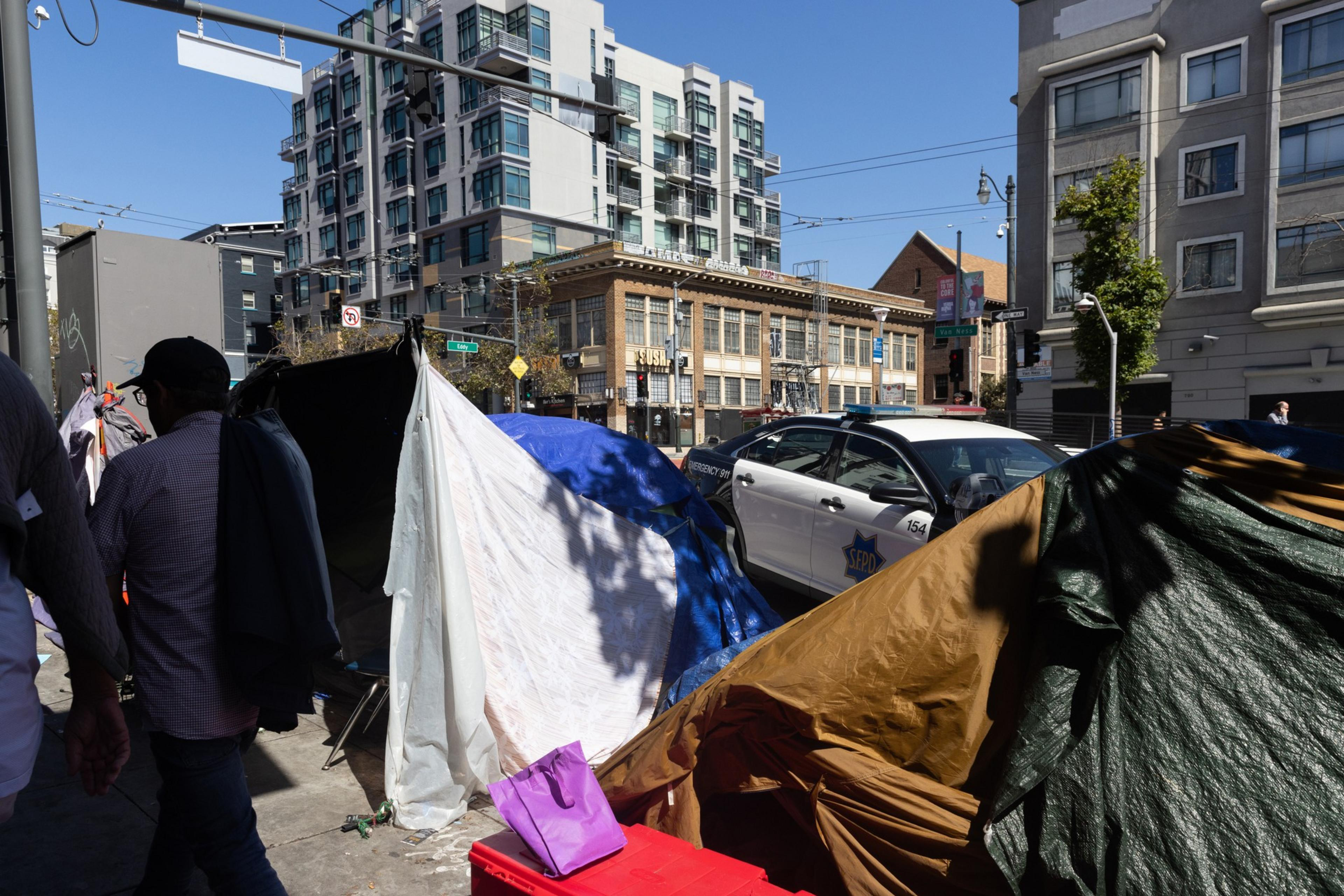 A police vehicle sits parked along Van Ness Avenue next to an encampment with tall buildings in the background.