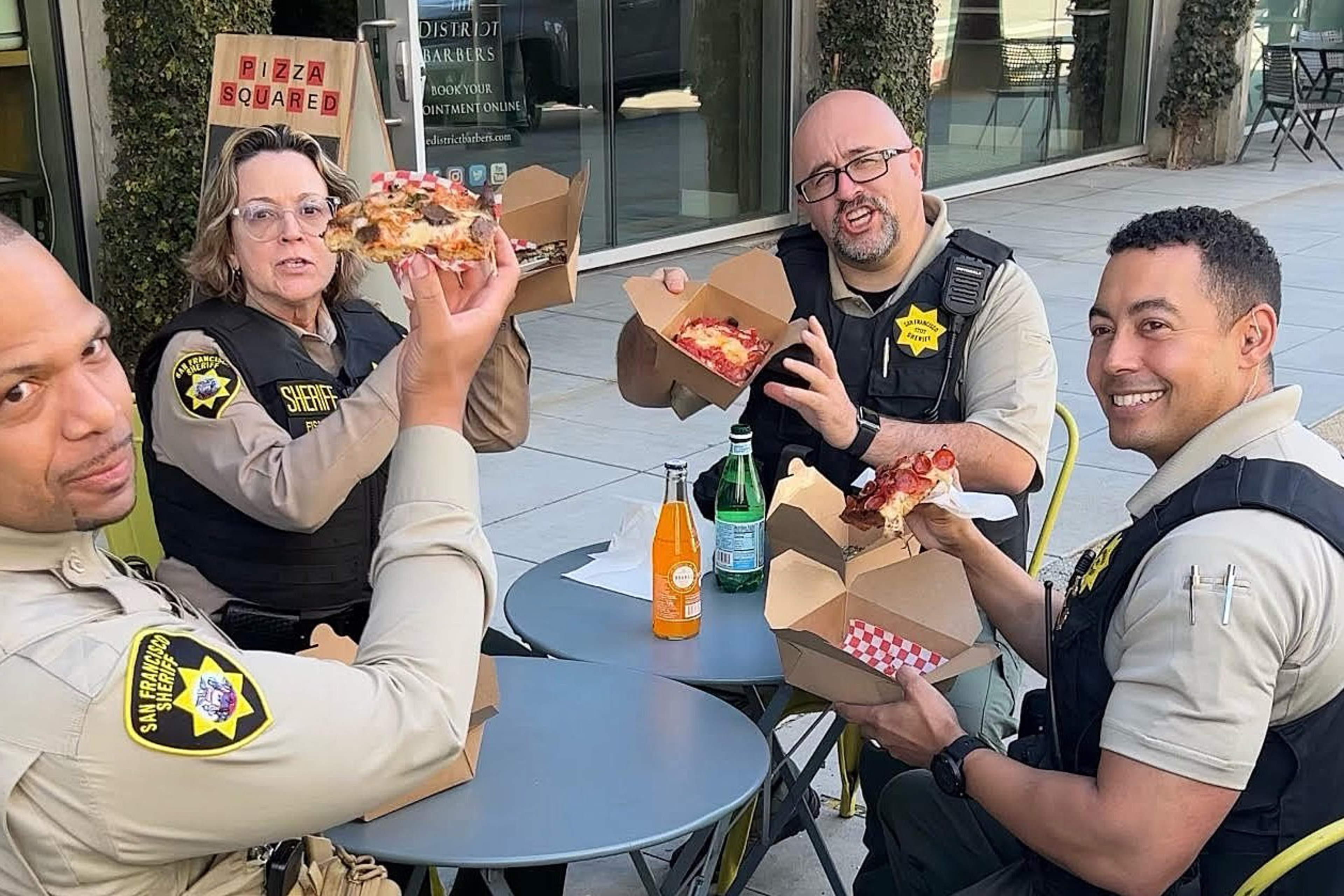 Four sheriff deputies wearing uniforms and vests sit outdoors at small round tables, smiling and holding pizzas in takeout boxes.